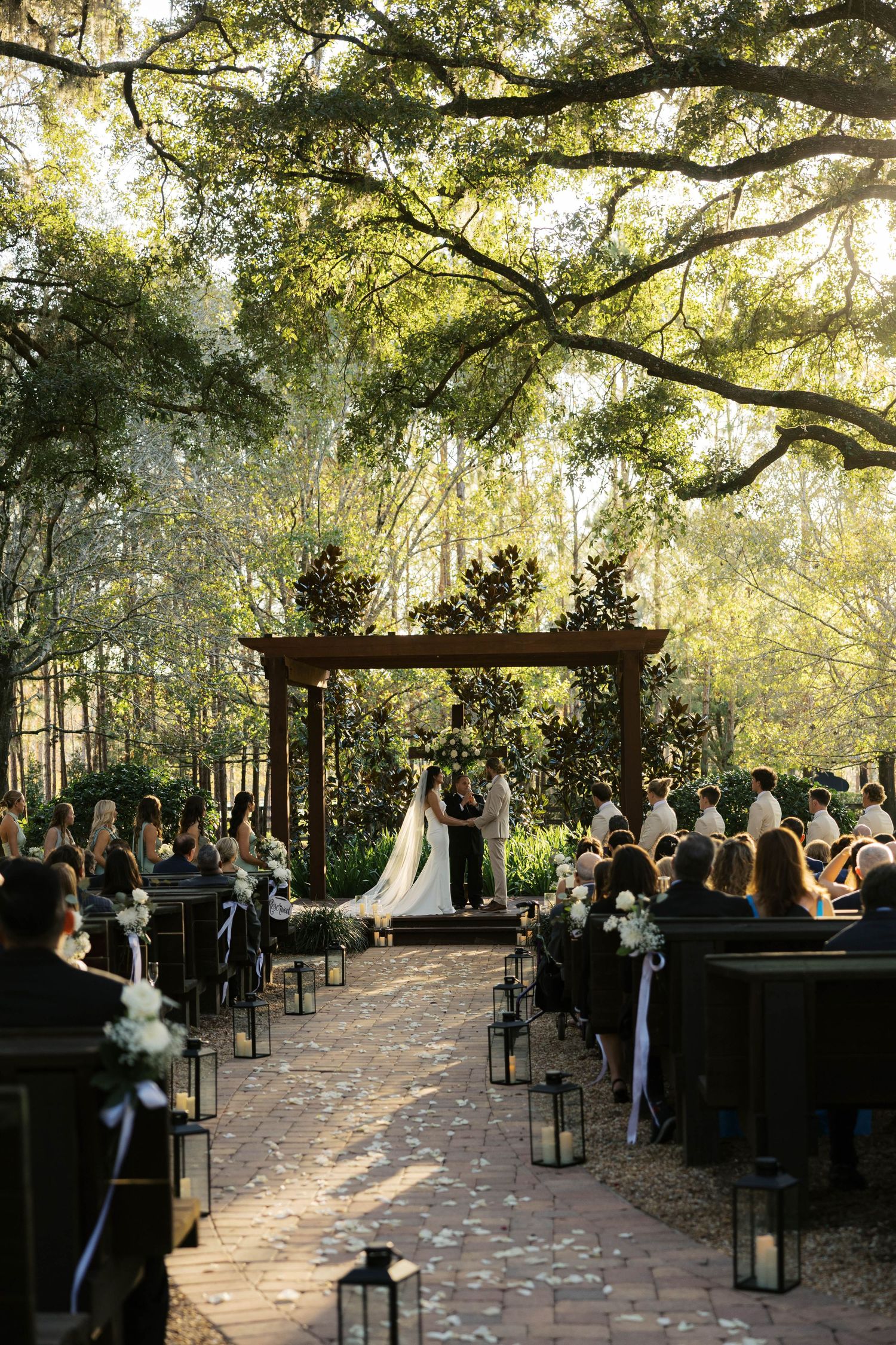 Outdoor wedding ceremony under oak trees with lantern-lined aisle.