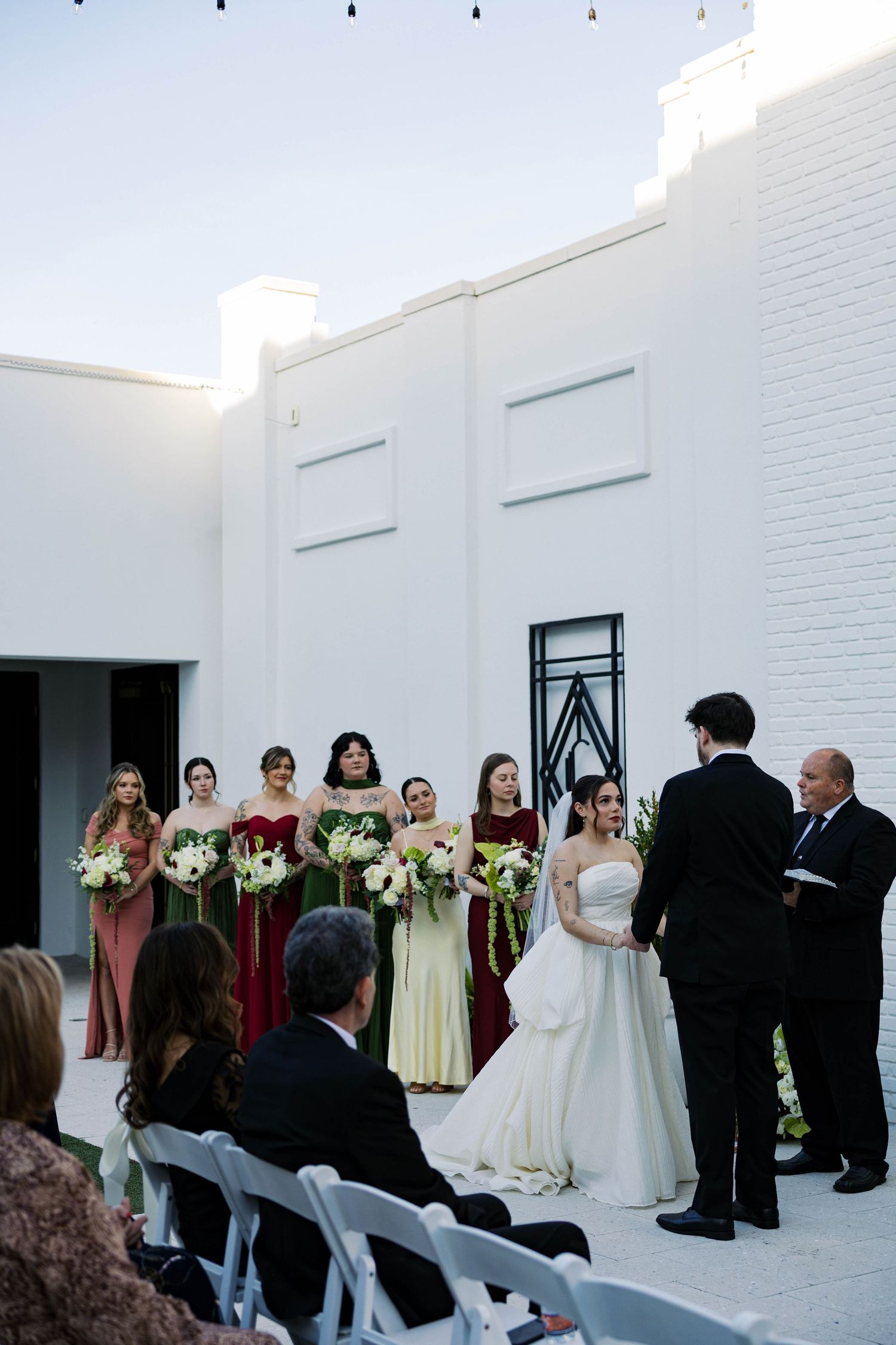 Wedding party lines up against white brick wall during outdoor ceremony with string lights overhead.