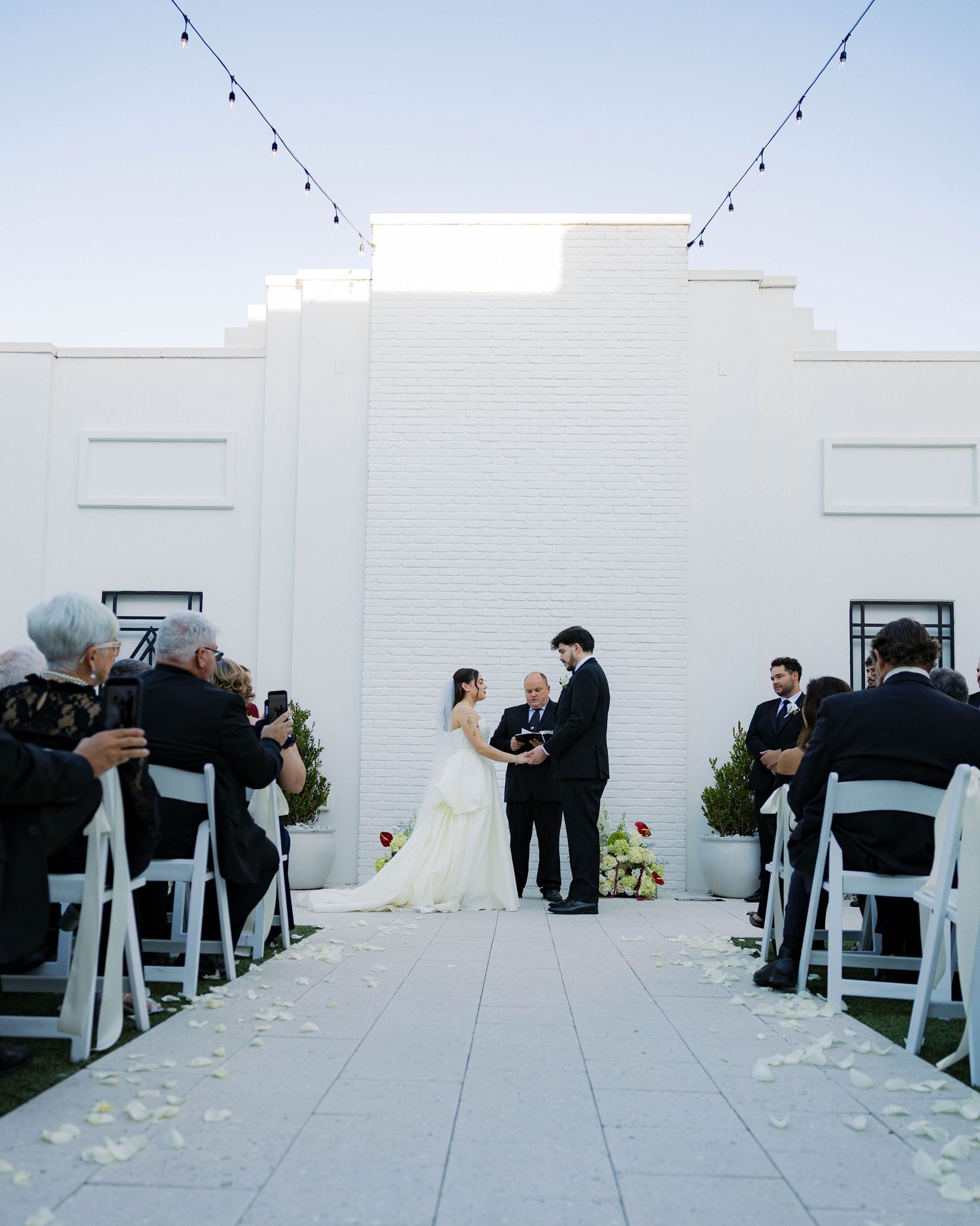 White chairs line outdoor wedding aisle with string lights and white brick backdrop.
