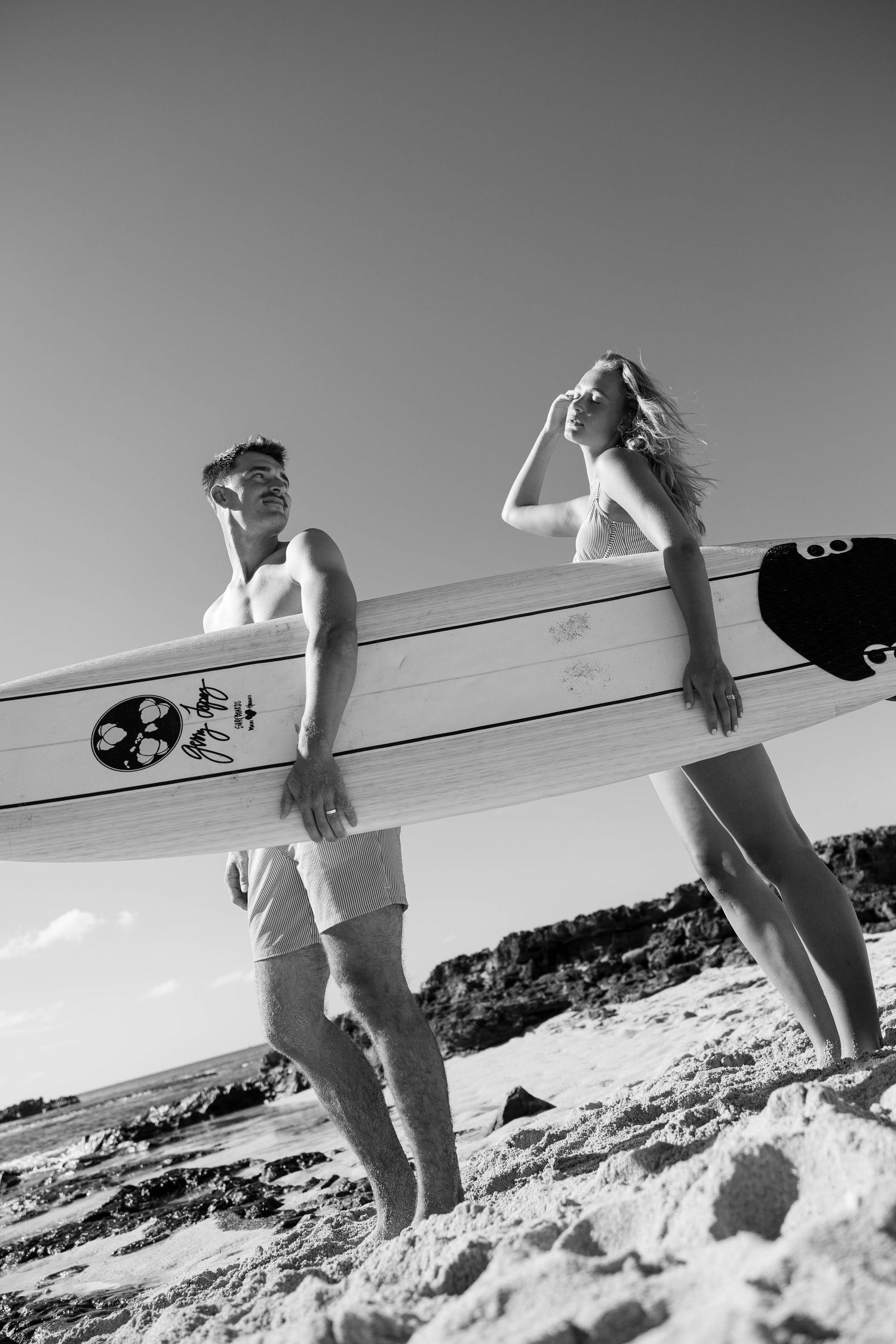 Black and white silhouettes carrying surfboards along a sandy beach against bright sky.