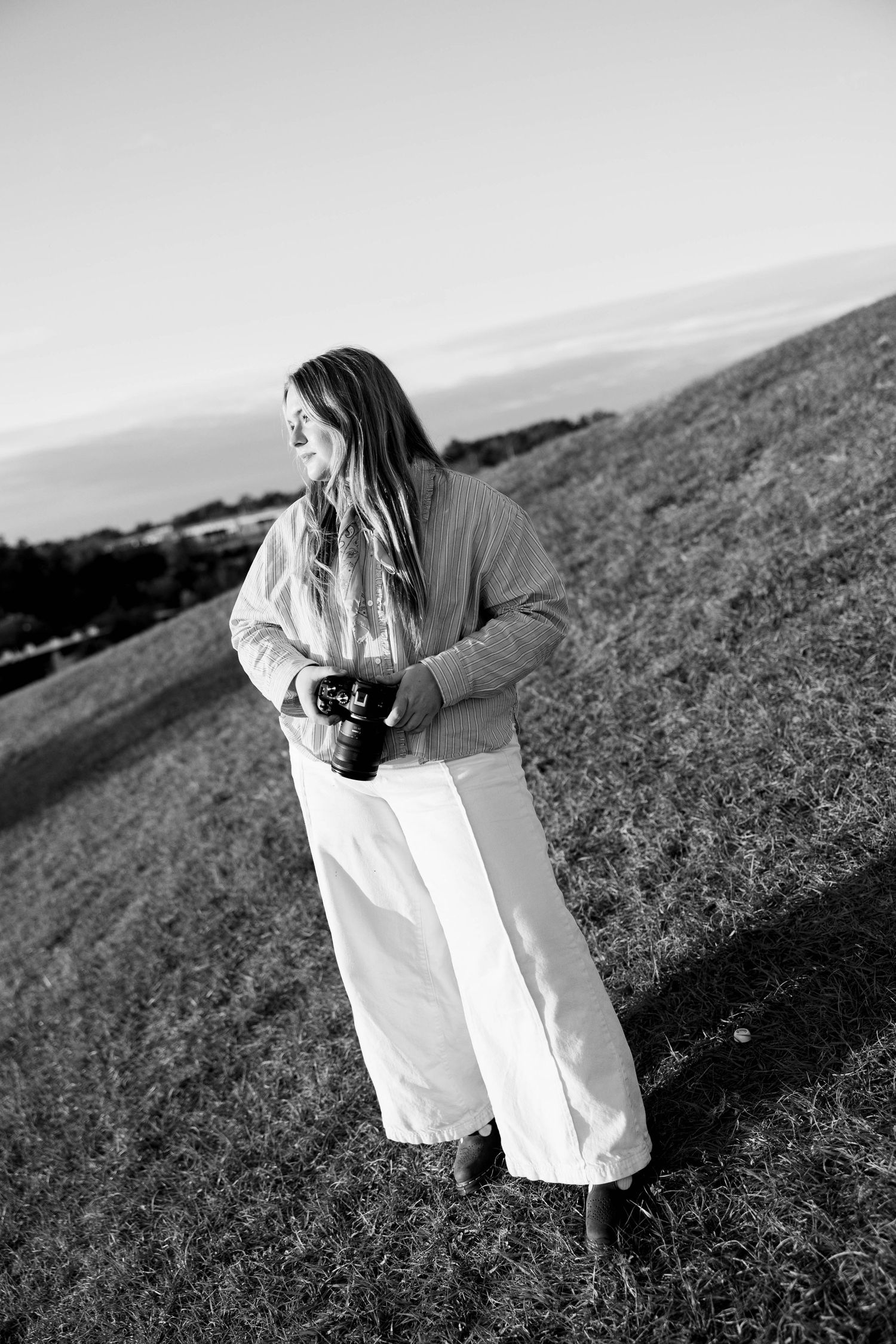 Black and white photo of someone in white pants and puffy jacket holding a camera on a hillside overlooking a landscape.
