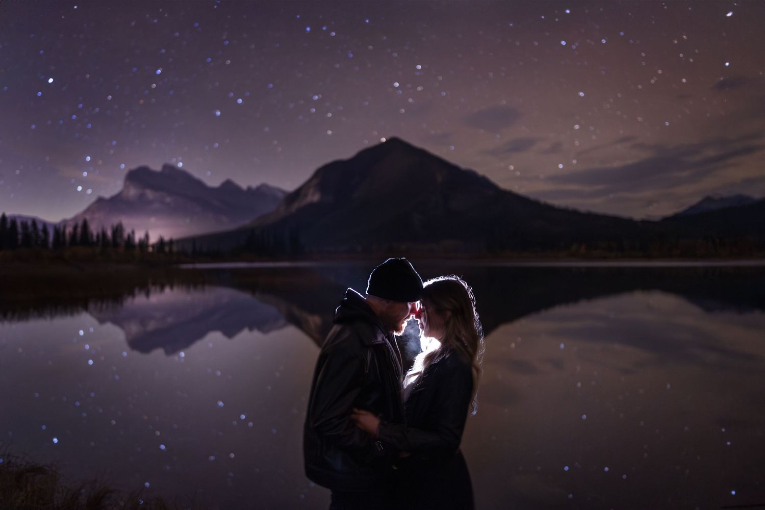 Silhouetted couple shares romantic moment under starry night sky beside mountain lake with reflective waters of Vermillion Lakes Banff