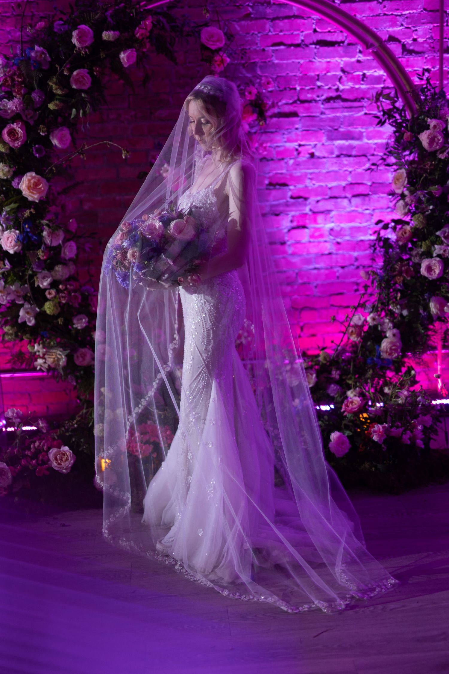 A bride in a lace wedding gown and long veil poses under a floral arch with purple lighting at Venue 308 Calgary.