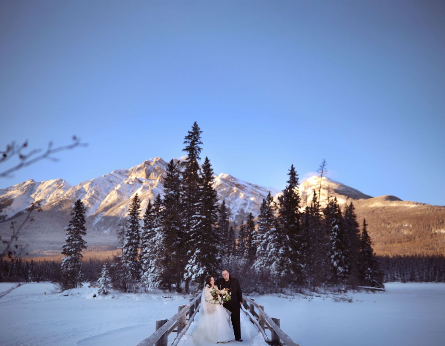 Snow-covered mountain peaks rise behind pyramid island lake with a bride and groom on their wedding day in jasper
