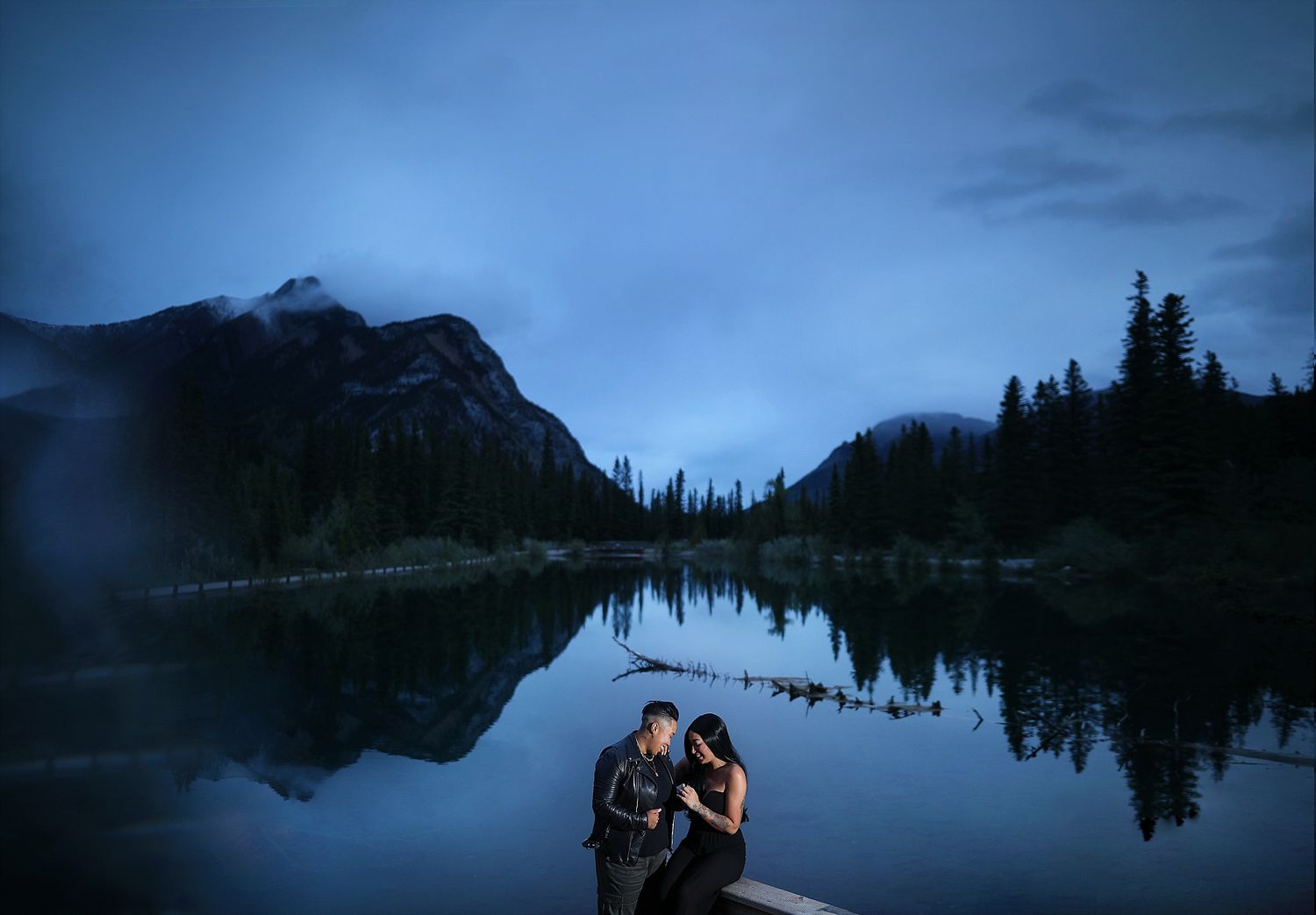 A silhouetted couple embraces by a mountain lake at twilight with perfect reflections in the waters of Mt Lorette Ponds Kananaskis Country