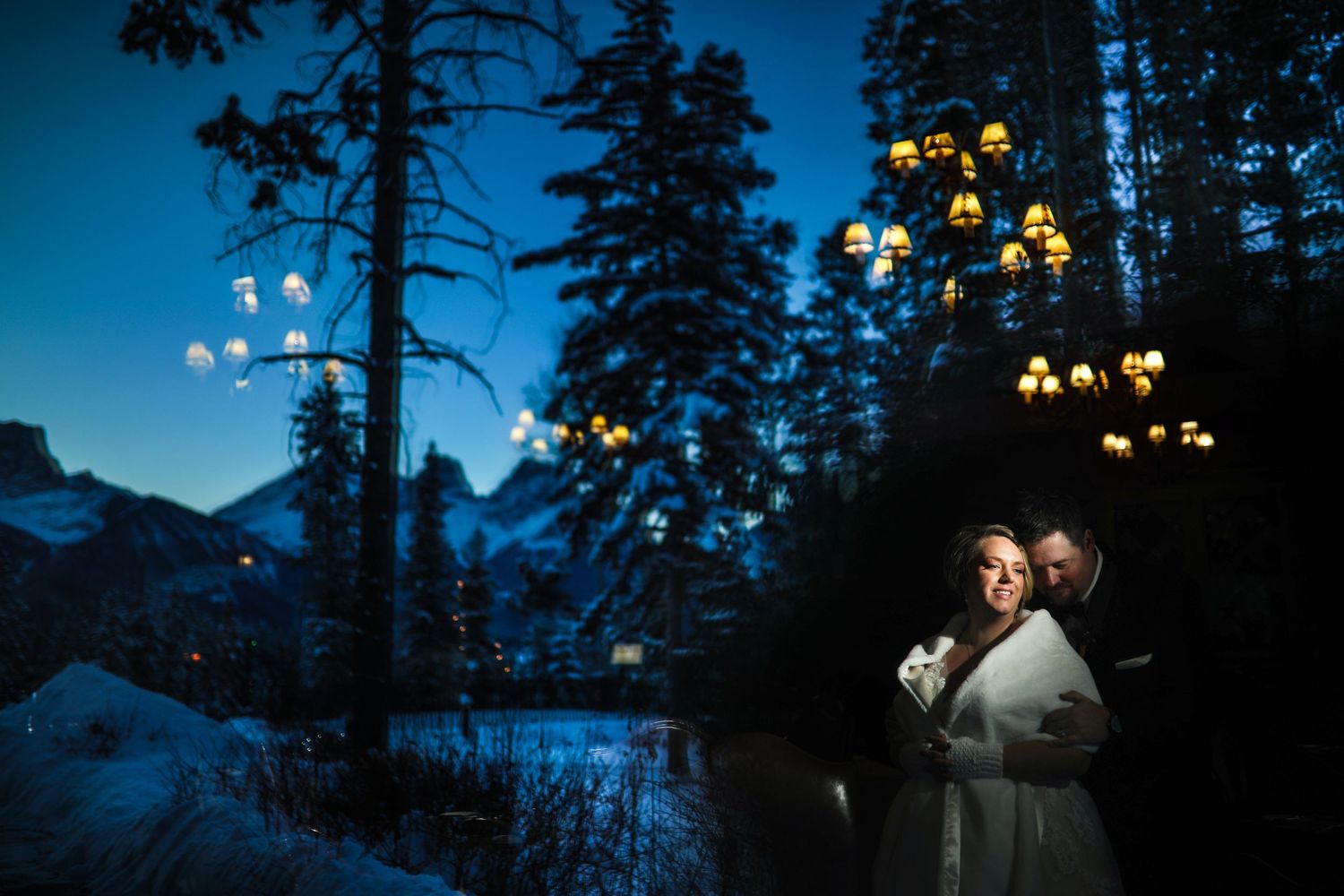 Couple in winter wedding attire illuminated by glowing lanterns against snowy mountain landscape at Silvertip Resort Canmore