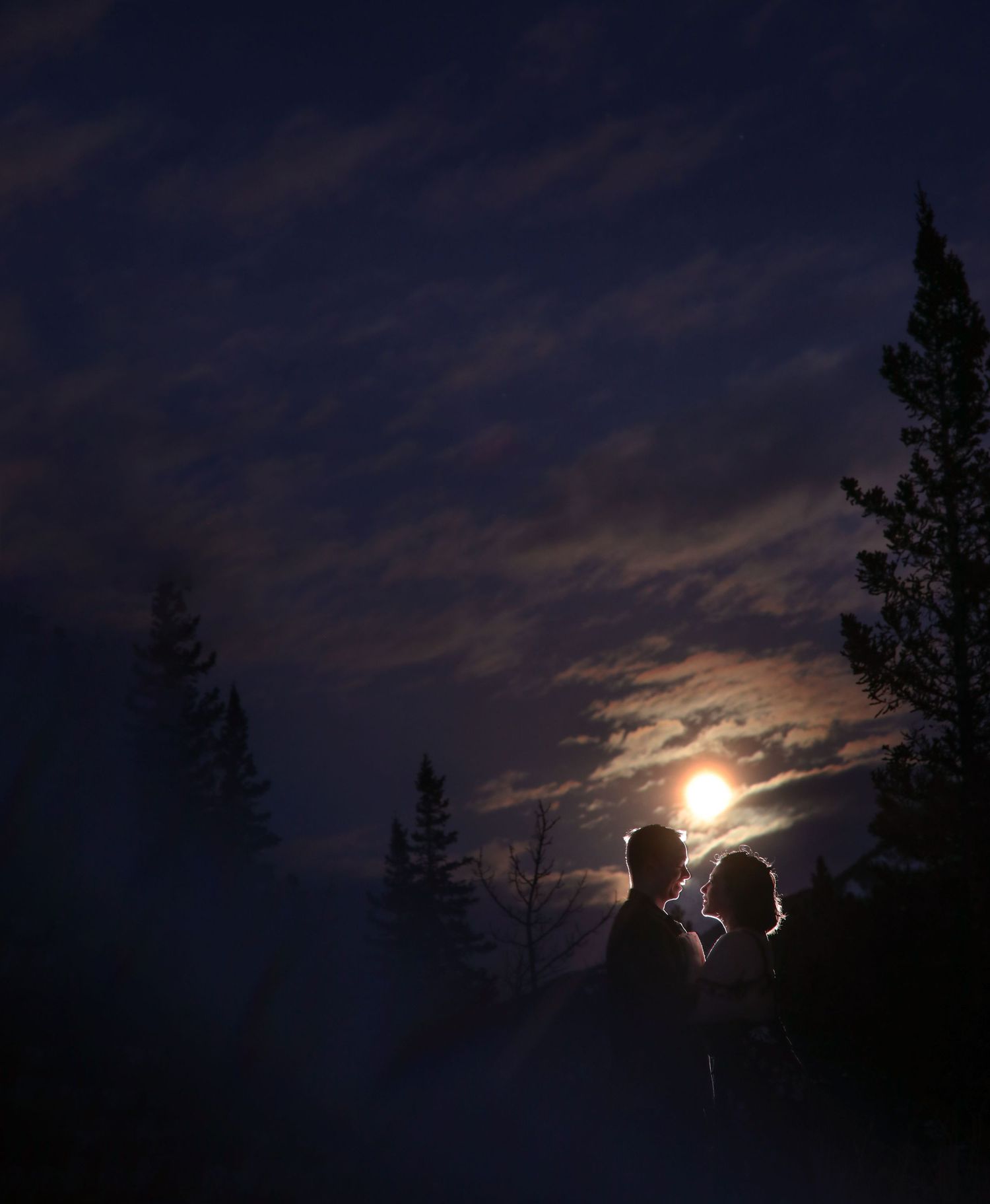 Couple share a romantic moment under a bright full moon with evergreen trees at Barrier Lake Kananaskis Country at night
