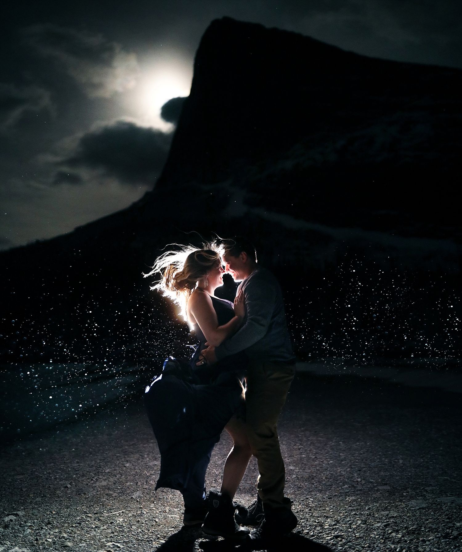 Silhouetted couple dancing together against a moody moonlit Hai Ling Peak in Canmore Alberta at night with the wind and rain around them.