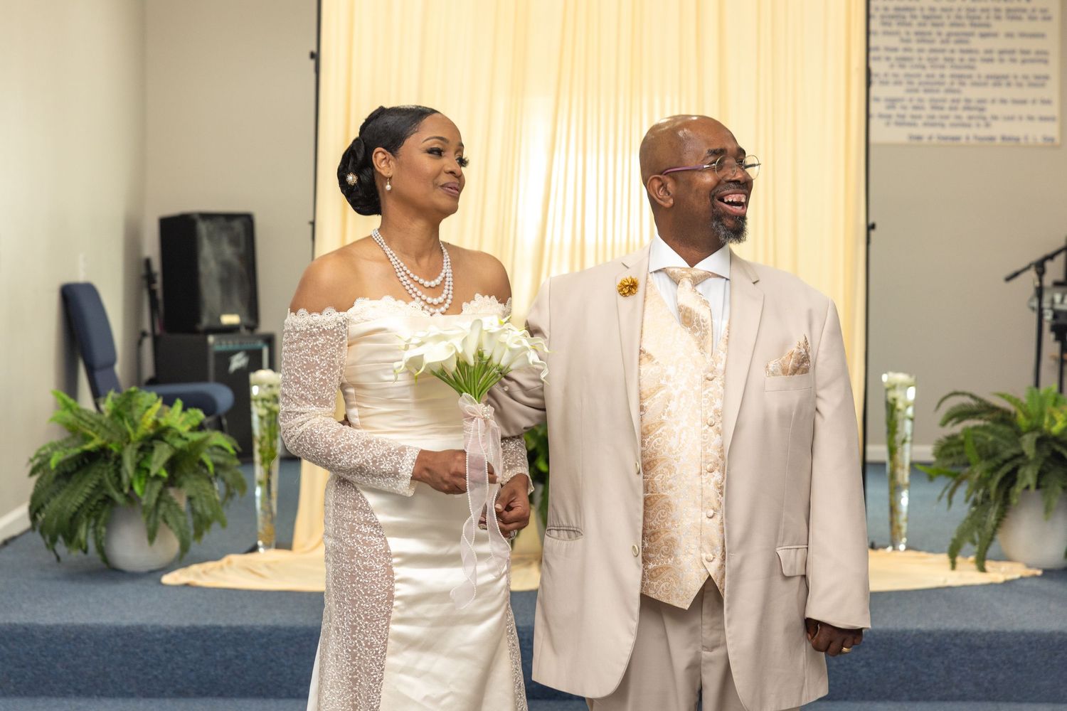 Bride in white gown and groom in beige suit holding hands at church wedding altar with floral decorations.
