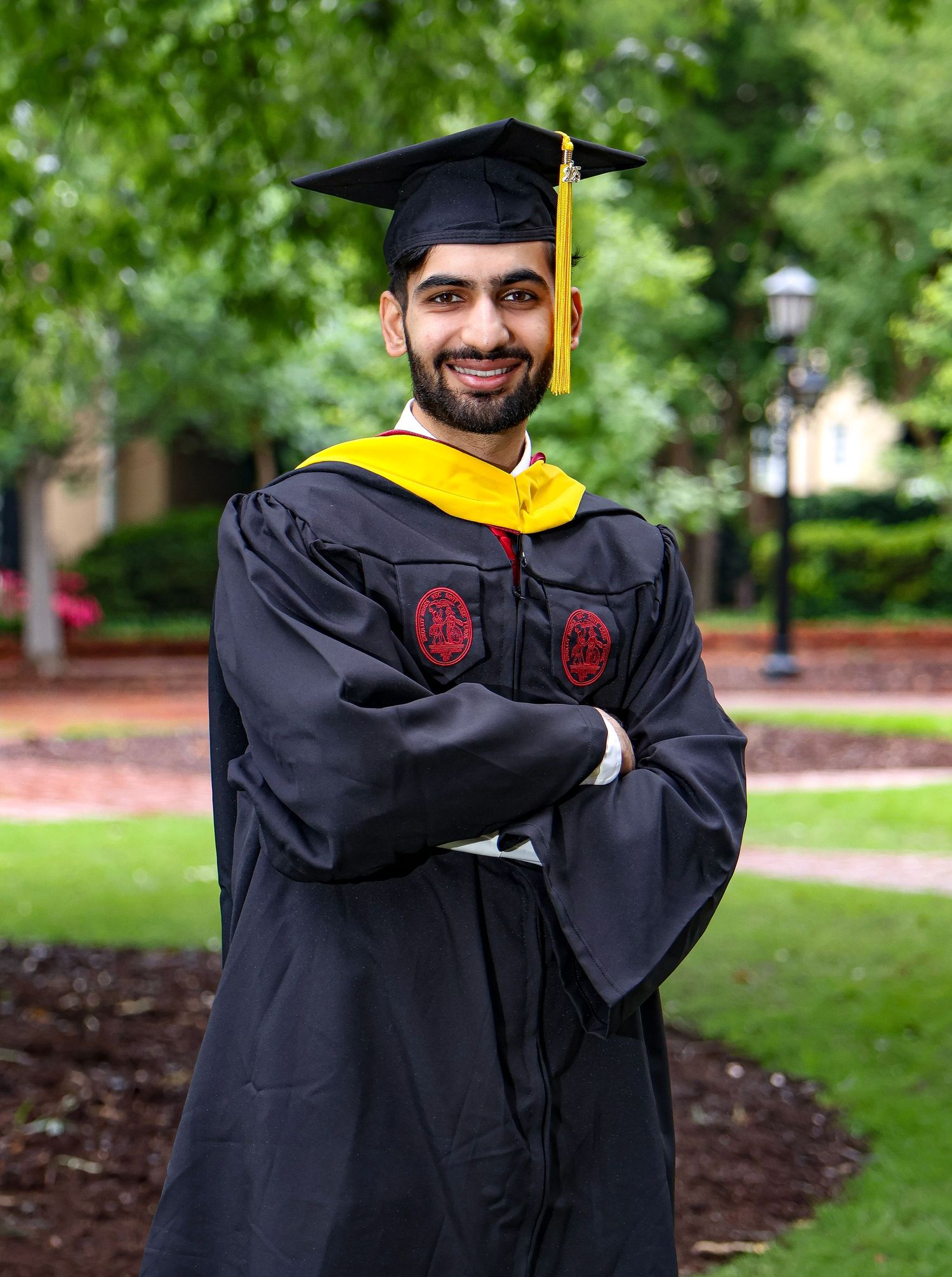 Graduate in academic regalia poses confidently with arms crossed in garden setting.
