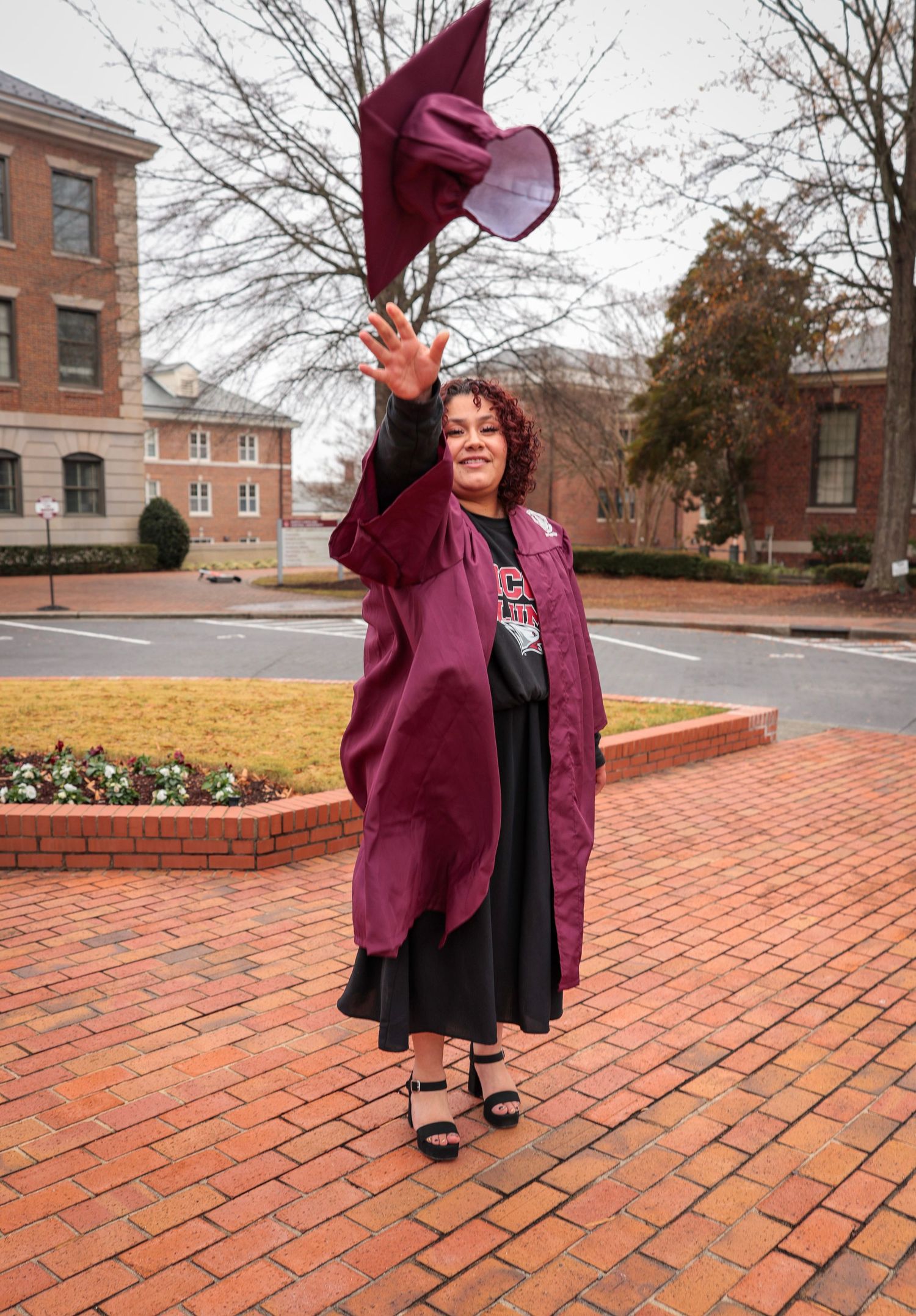 Graduate in maroon regalia poses playfully on brick courtyard.