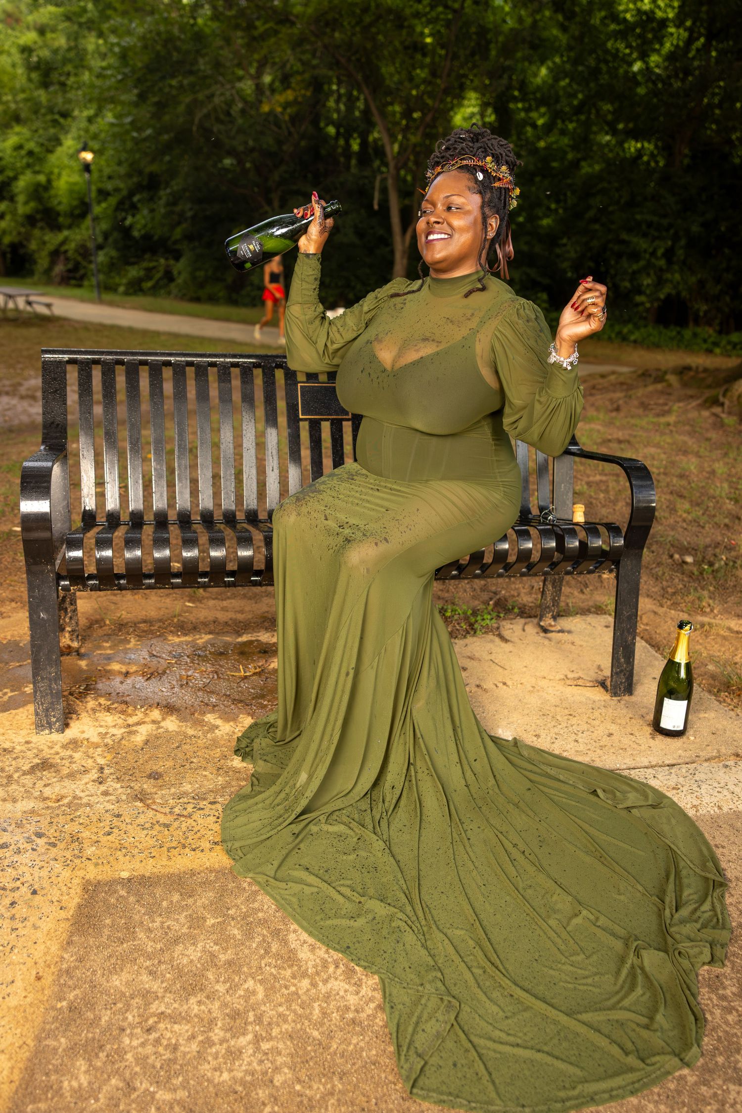 Woman in elegant olive green gown sitting on park bench holding champagne bottle, smiling joyfully outdoors.