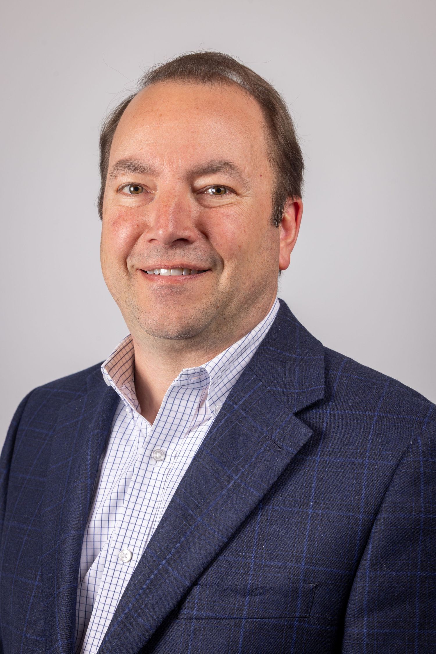 Professional headshot of a smiling middle-aged man in a navy plaid blazer and checkered shirt against a gray background.