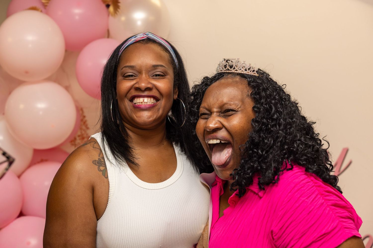 Two women laughing and posing at a pink balloon party, one wearing a tiara and bright pink top.