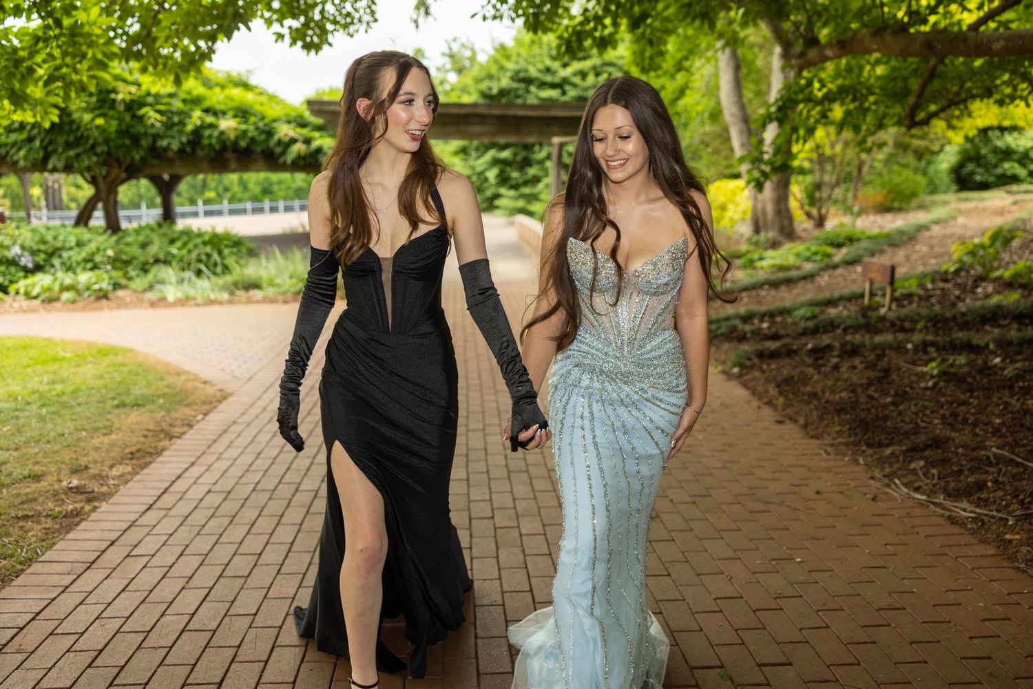 Two young women in elegant prom gowns walking hand in hand on a brick path in a lush garden.
