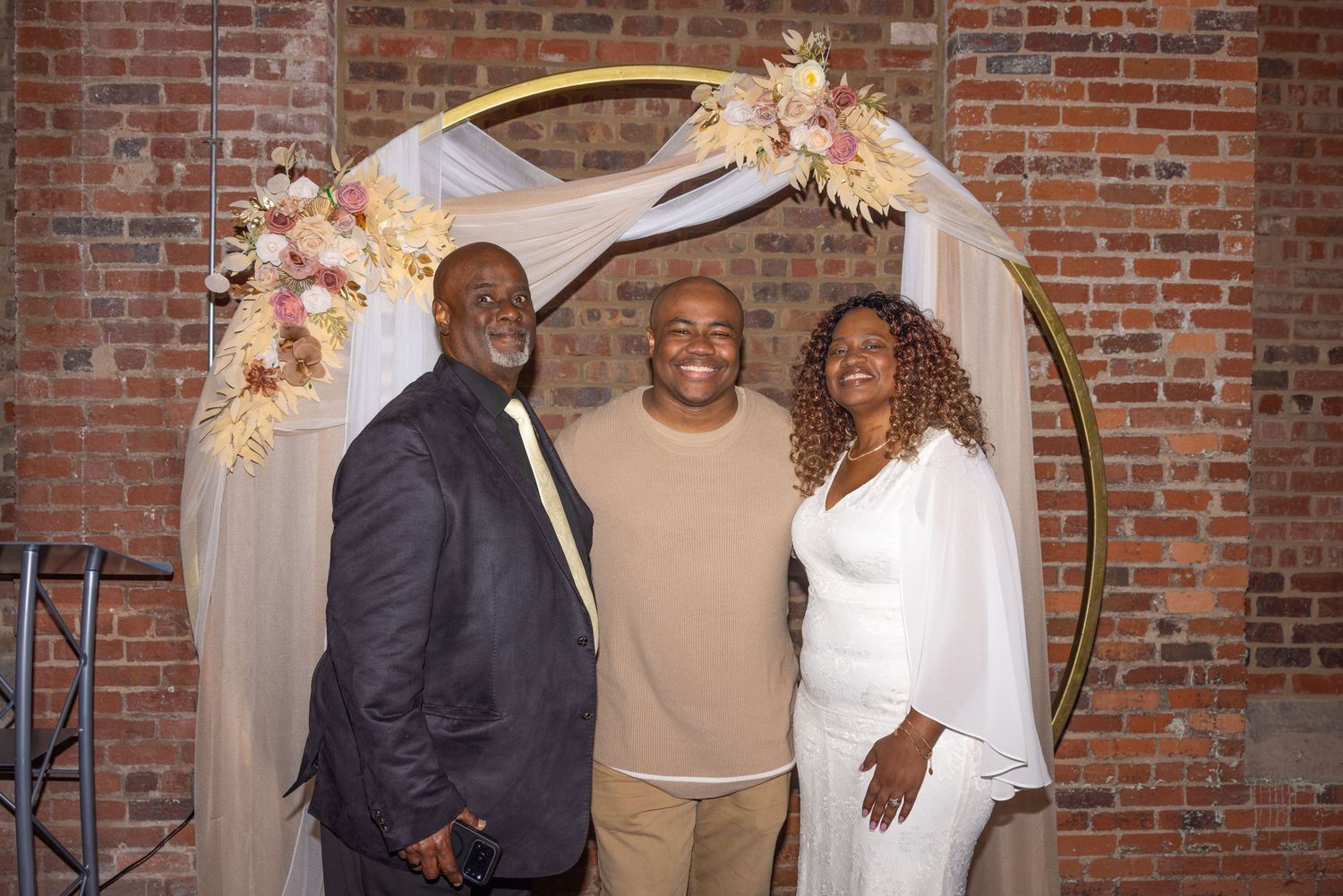 Three people smiling together in front of a gold circular arch with floral decorations at an elegant event venue.
