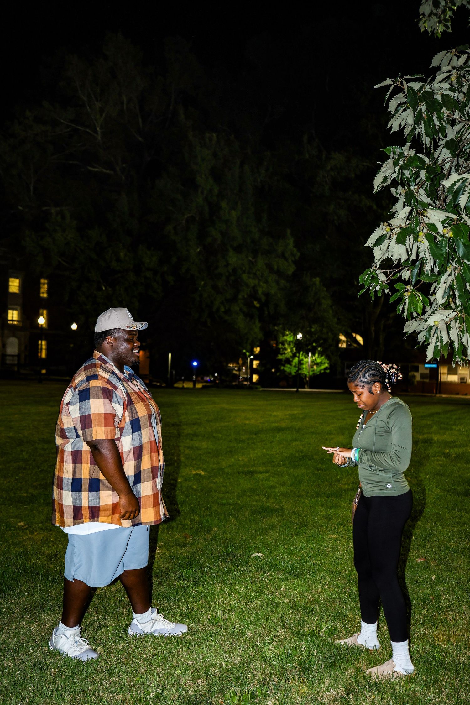 A couple stands on a grassy lawn at night with buildings and trees visible in the background.