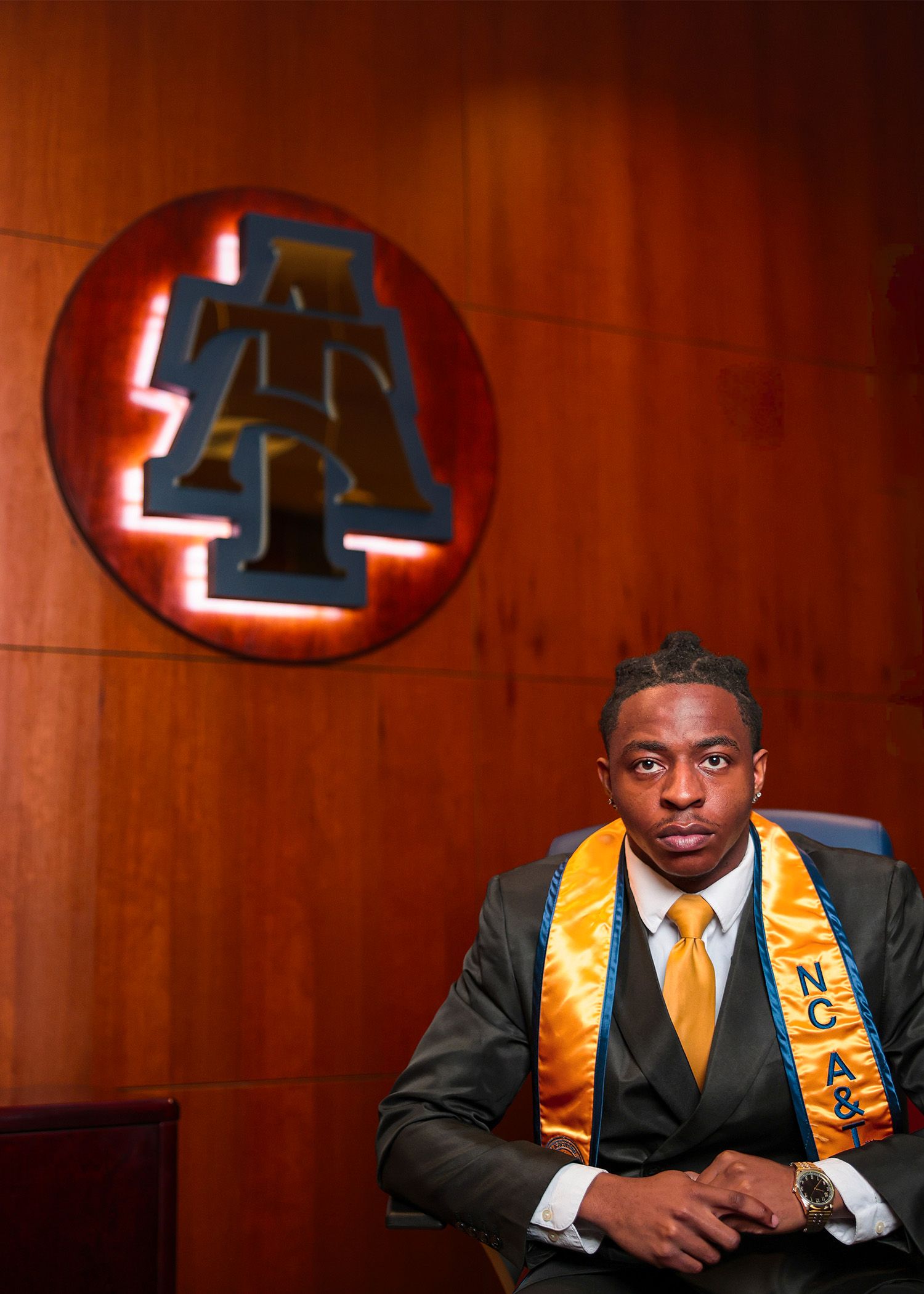 Graduate in formal attire poses beneath illuminated numbers on red wall.