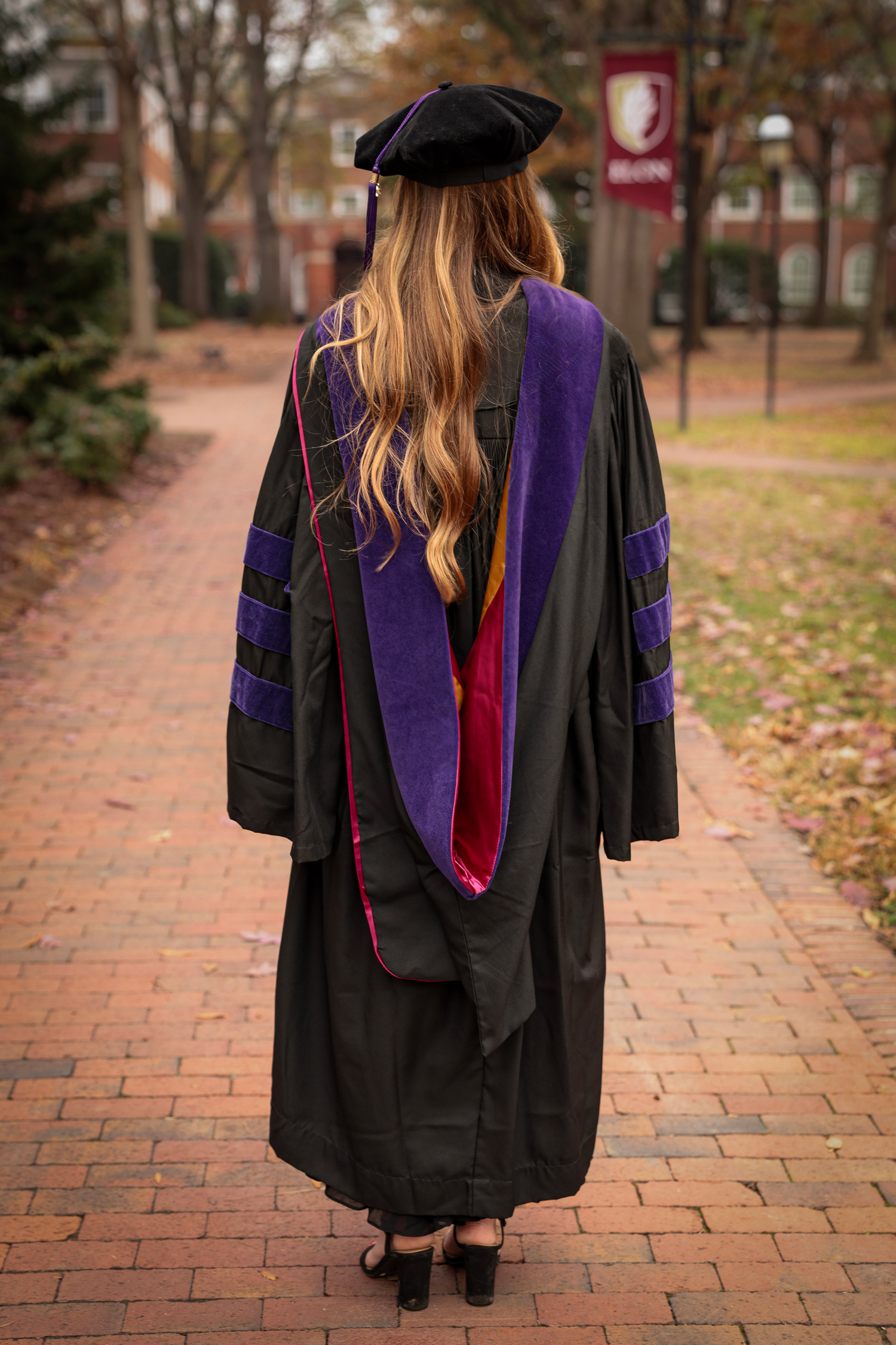 Back view of graduate in academic regalia showing purple hood and black cap on campus walkway.