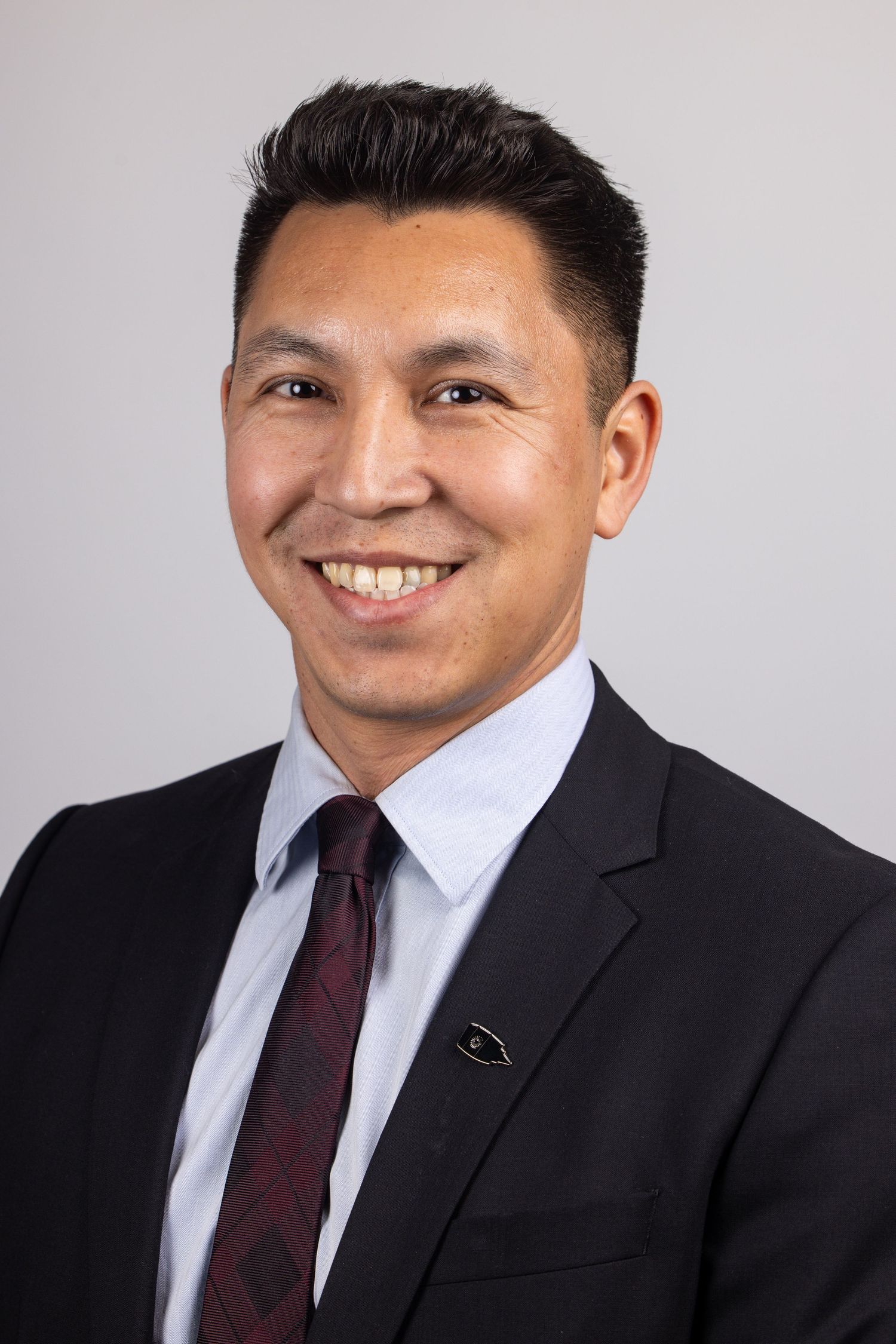 Professional headshot of a smiling man in a dark suit and burgundy tie against a gray background.