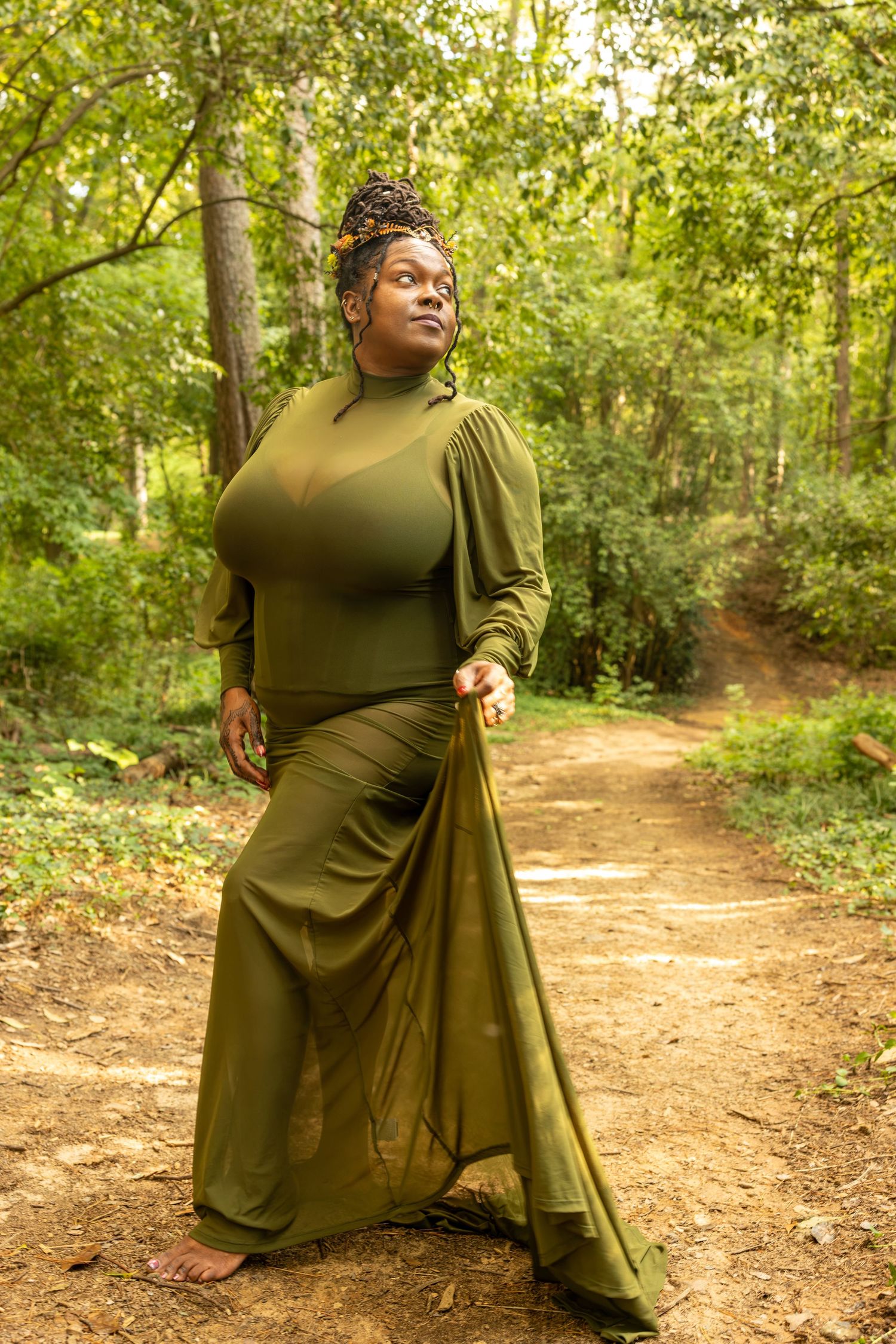 Woman in elegant olive green gown poses barefoot on forest path surrounded by lush green trees.