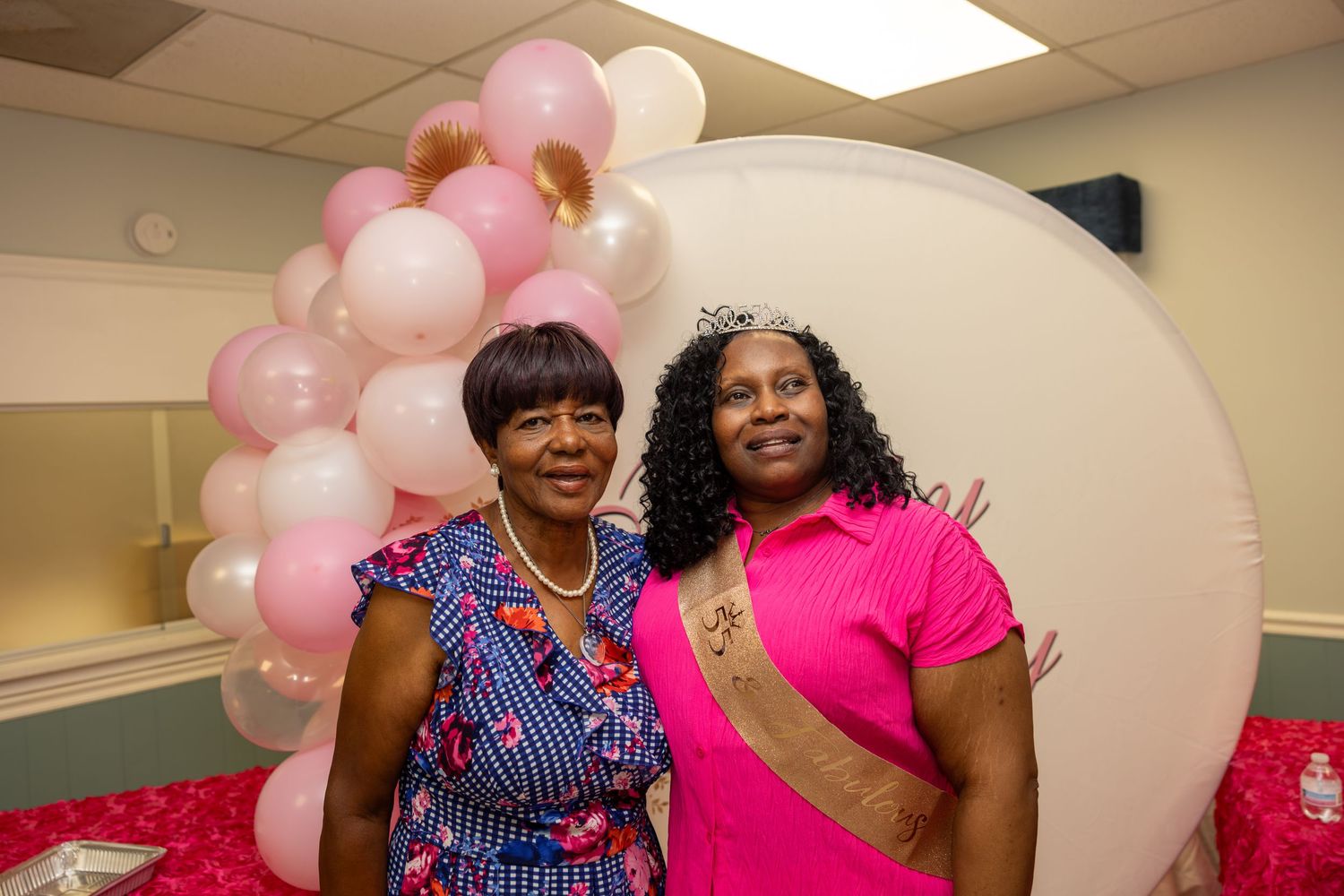 Two women posing together at a pink-themed celebration with balloon arch and decorations.