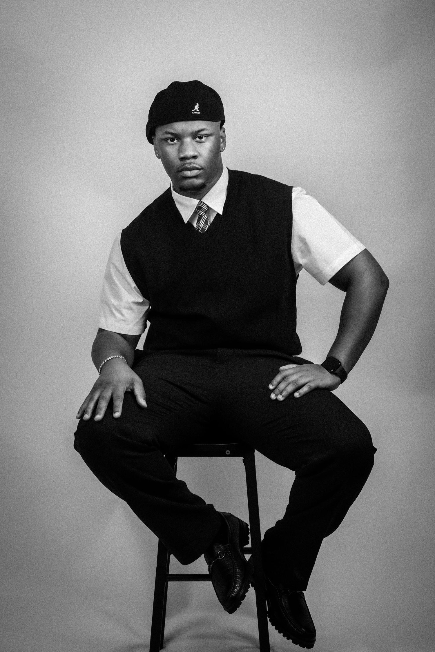 Black and white portrait of a young man in a vest, tie, and cap sitting confidently on a stool.