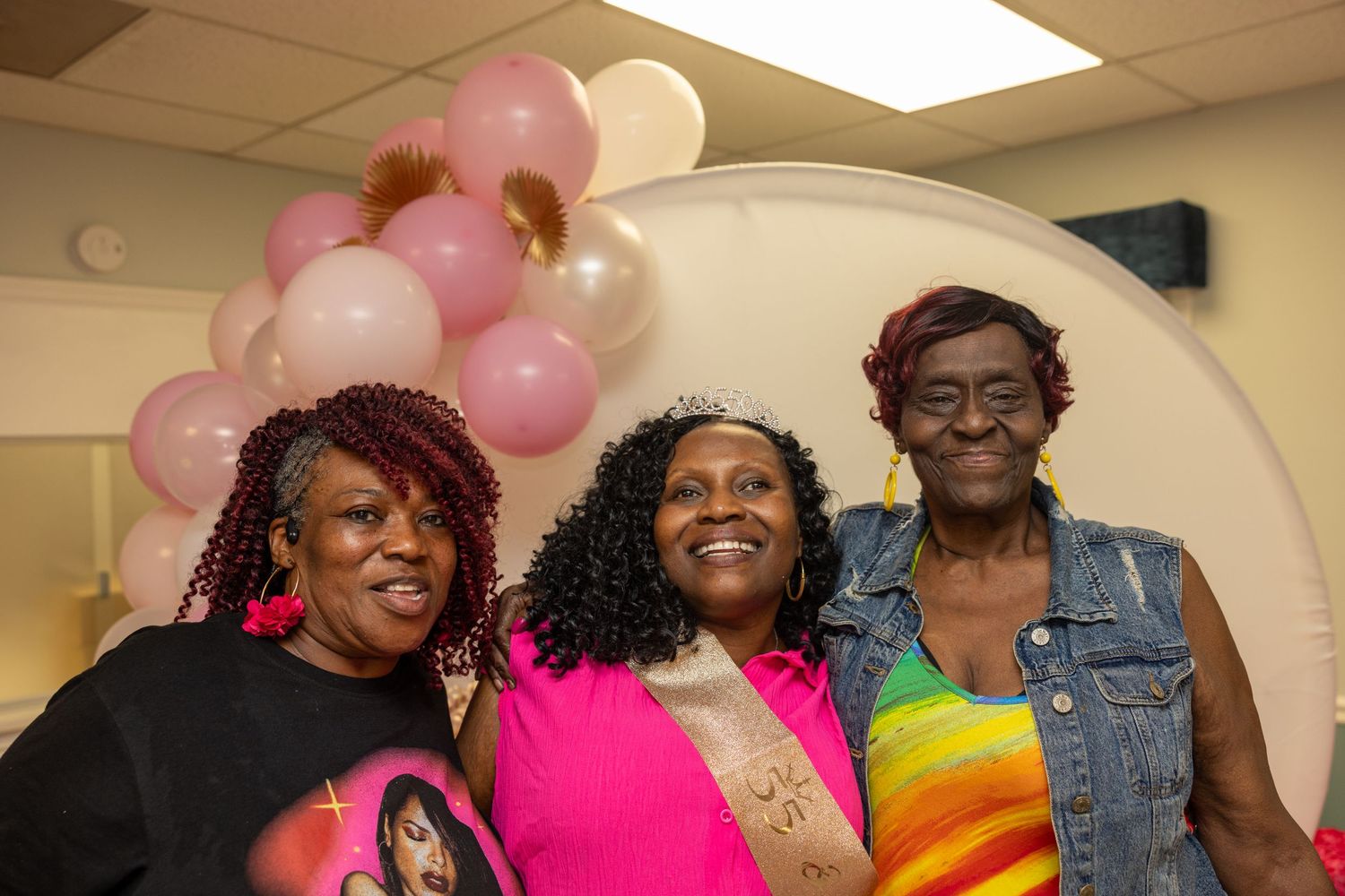 Three women smiling together at a celebration event with pink and white balloon decorations.