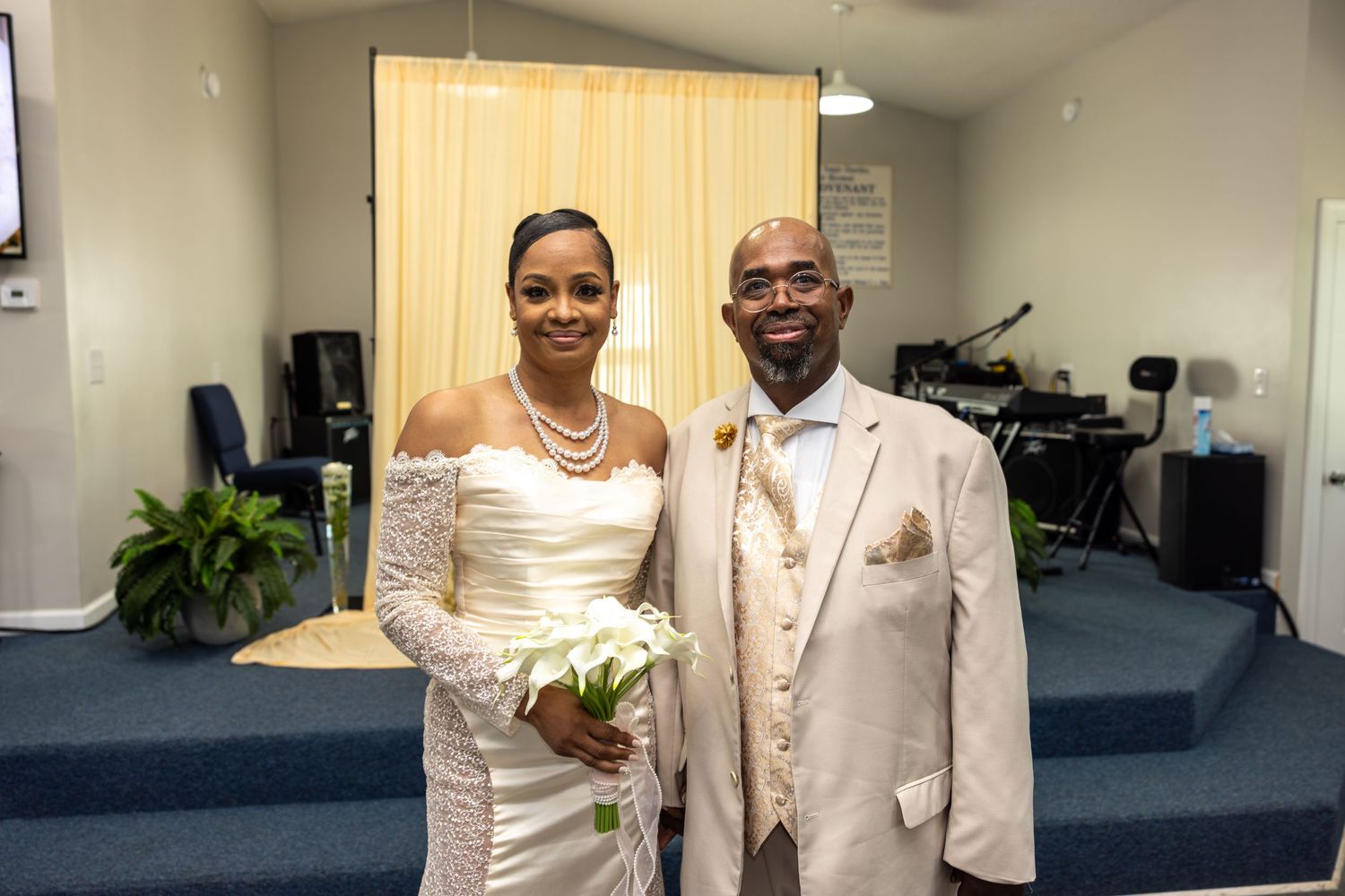 Bride and groom posing together in a church, wearing elegant white and cream wedding attire with pearl accessories.