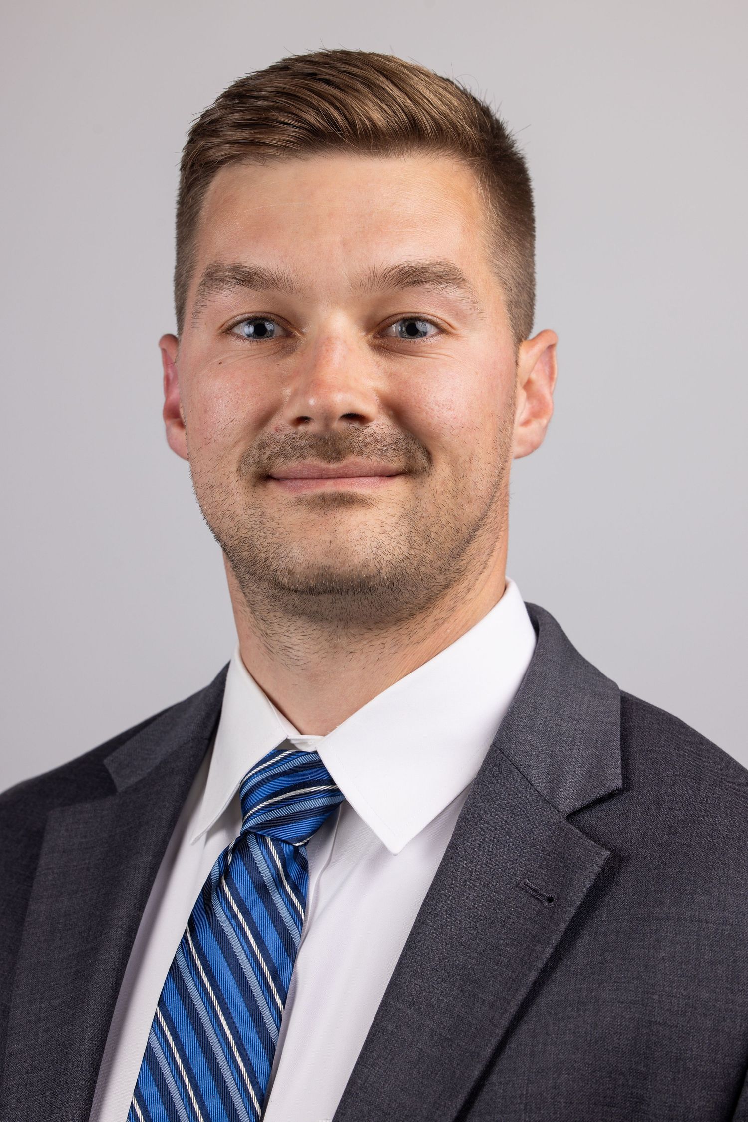 Professional headshot of a young man in a gray suit with a blue striped tie and white shirt against a gray background.