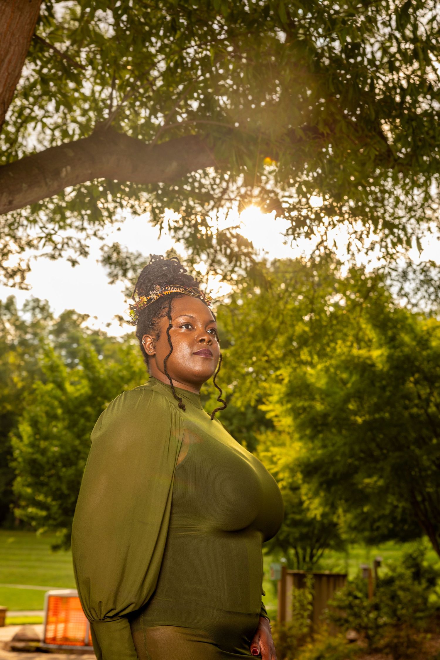 Woman in olive green dress poses under sunlit tree in lush park, golden hour light glowing behind her.