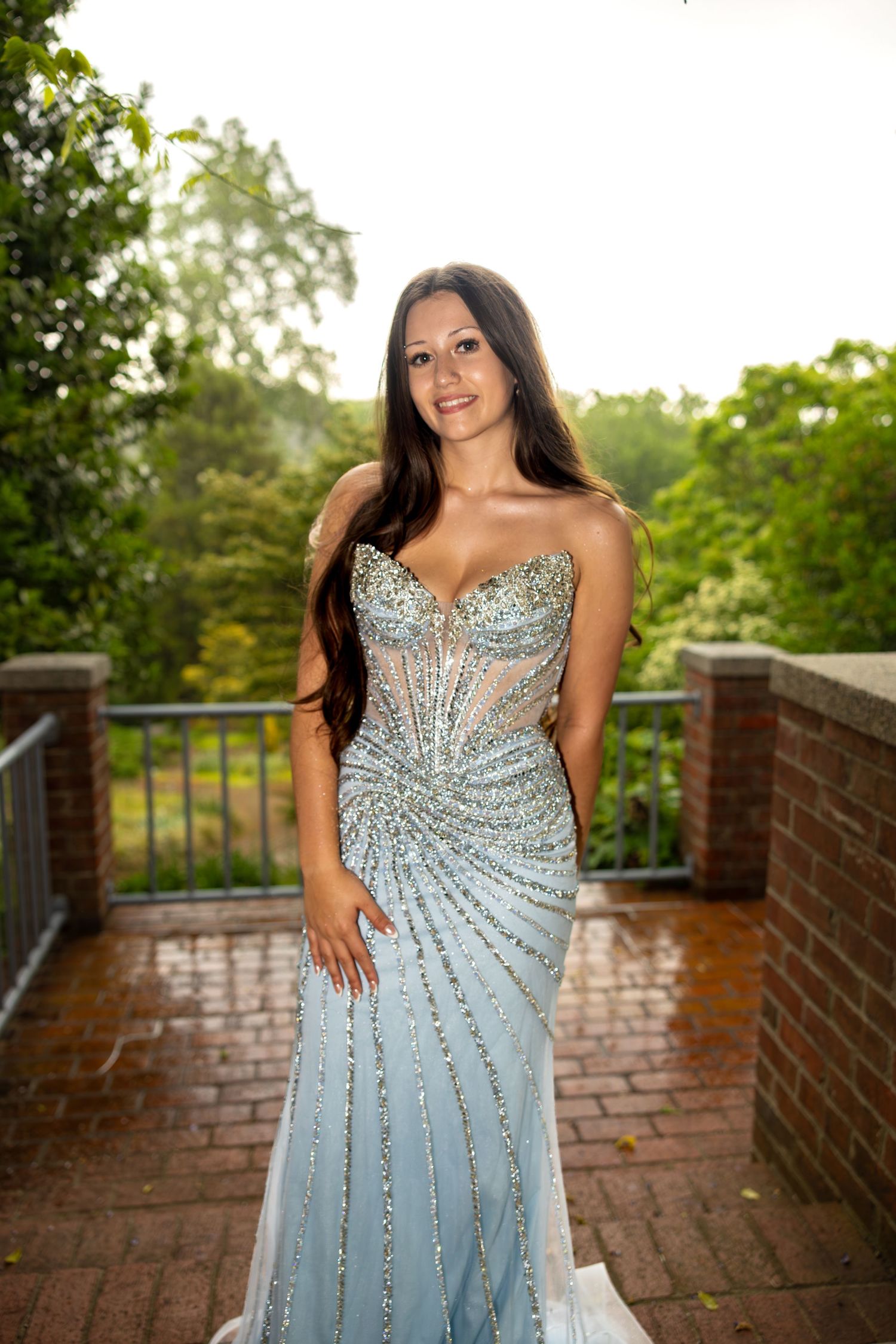 Young woman in a beaded strapless light blue prom gown posing on an outdoor terrace surrounded by lush greenery.