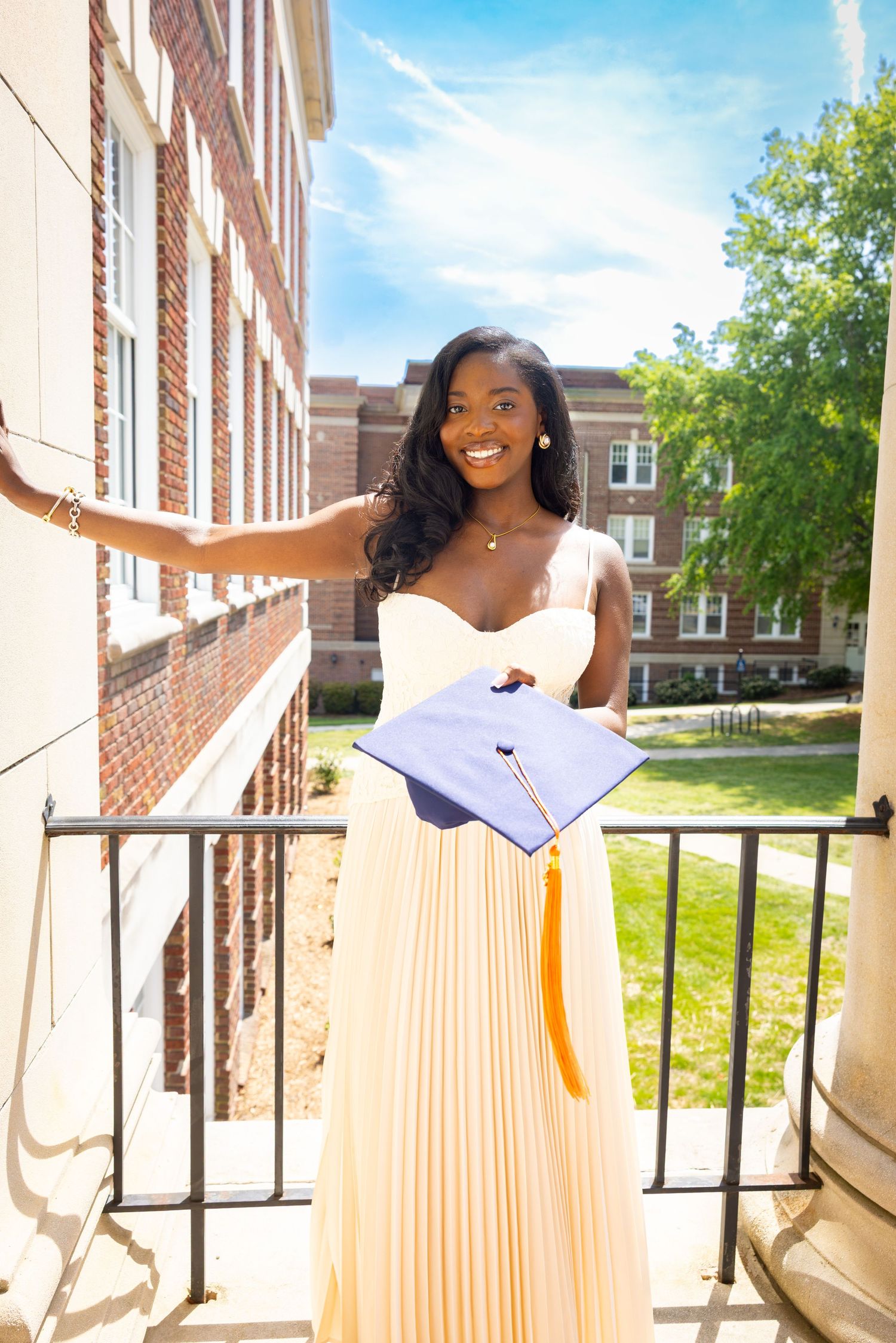 Smiling graduate in yellow dress holds blue cap with gold tassel on sunny campus steps.