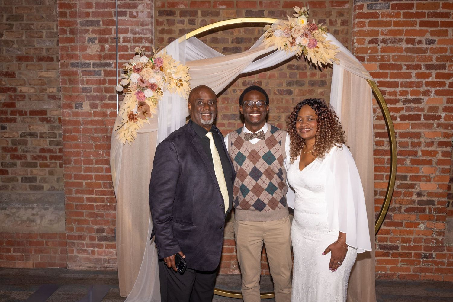 Three people smiling together under a gold circular arch with floral decor against a brick wall backdrop.