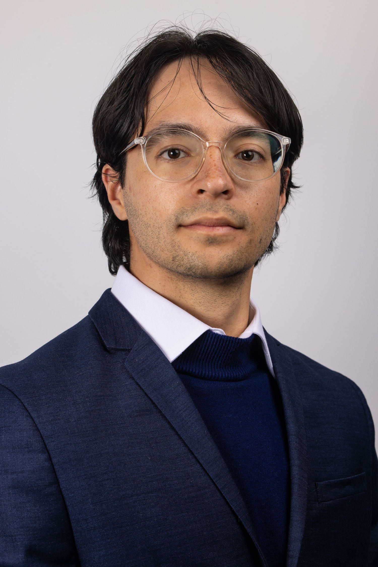 Professional headshot of a young man wearing glasses and a navy blue blazer against a gray background.