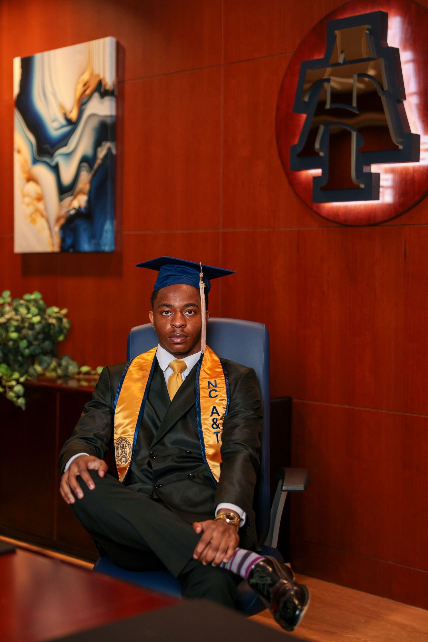 Graduate in formal black suit and gold stoles poses in elegant wood-paneled room.
