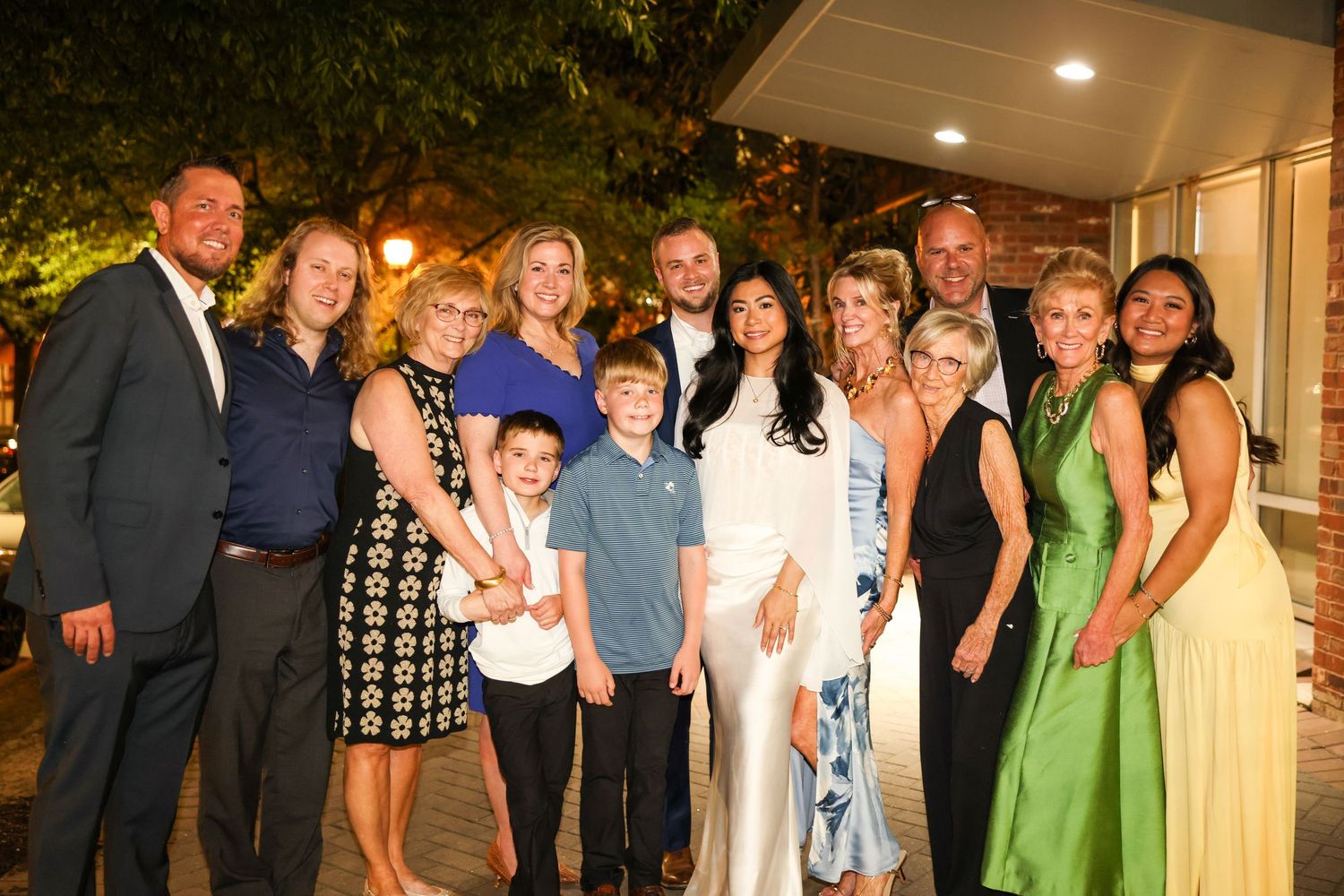 Group of smiling adults and children posing together outdoors at a formal evening event, dressed in elegant attire.