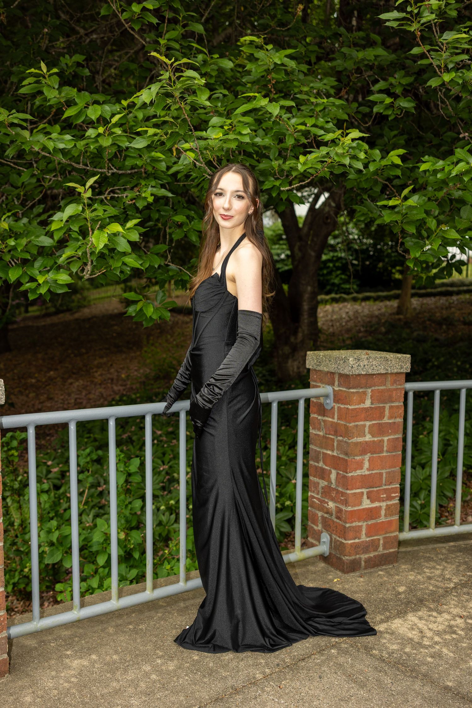 Young woman in elegant black satin gown with long gloves poses gracefully outdoors near brick pillar and metal railing.