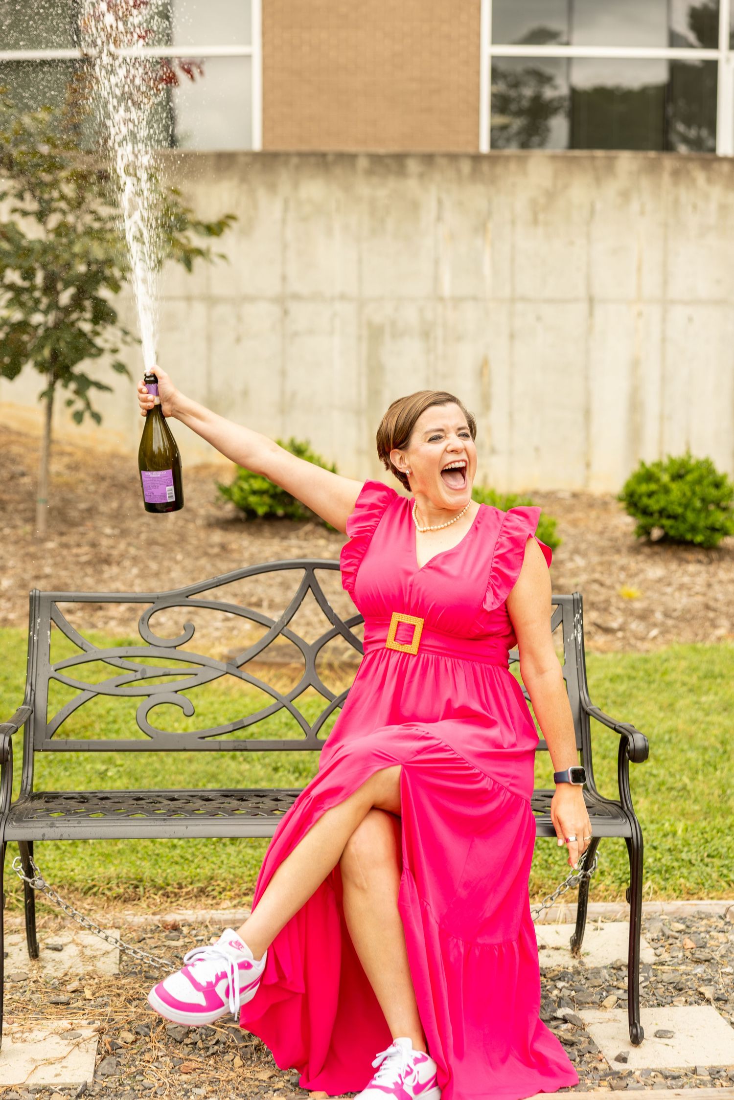 Woman in pink dress celebrates on bench, popping champagne bottle outdoors in joyful pose.