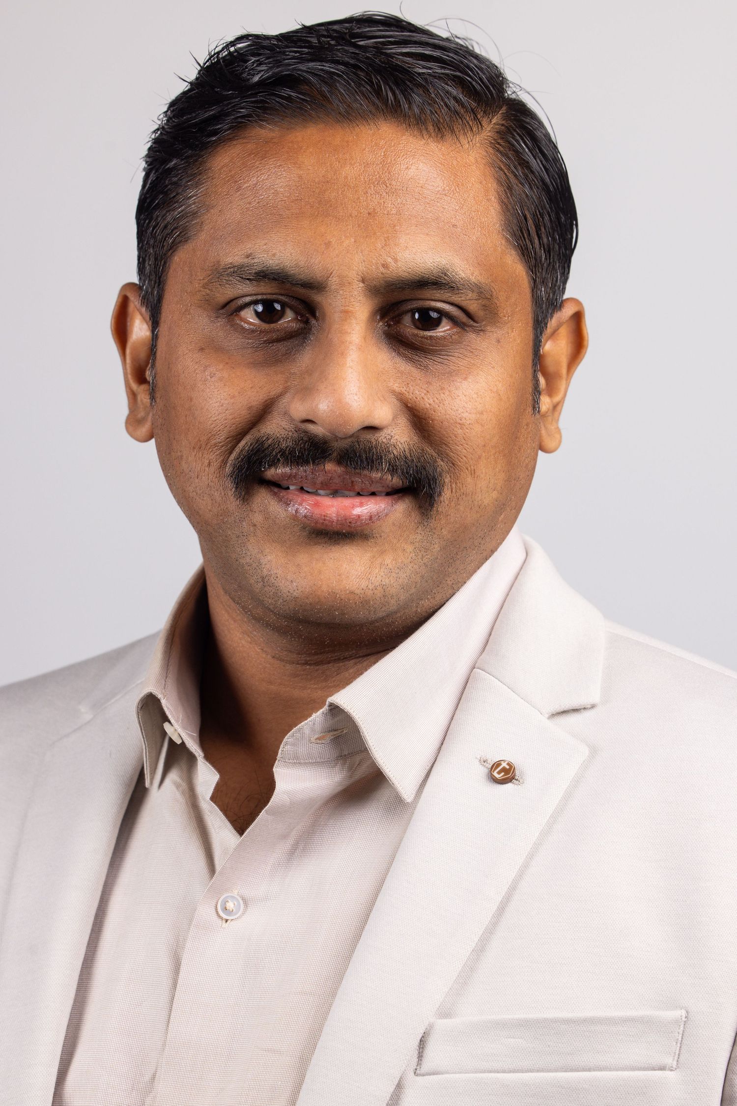 Professional headshot of a smiling South Asian man with mustache wearing a light beige blazer and shirt against gray background.