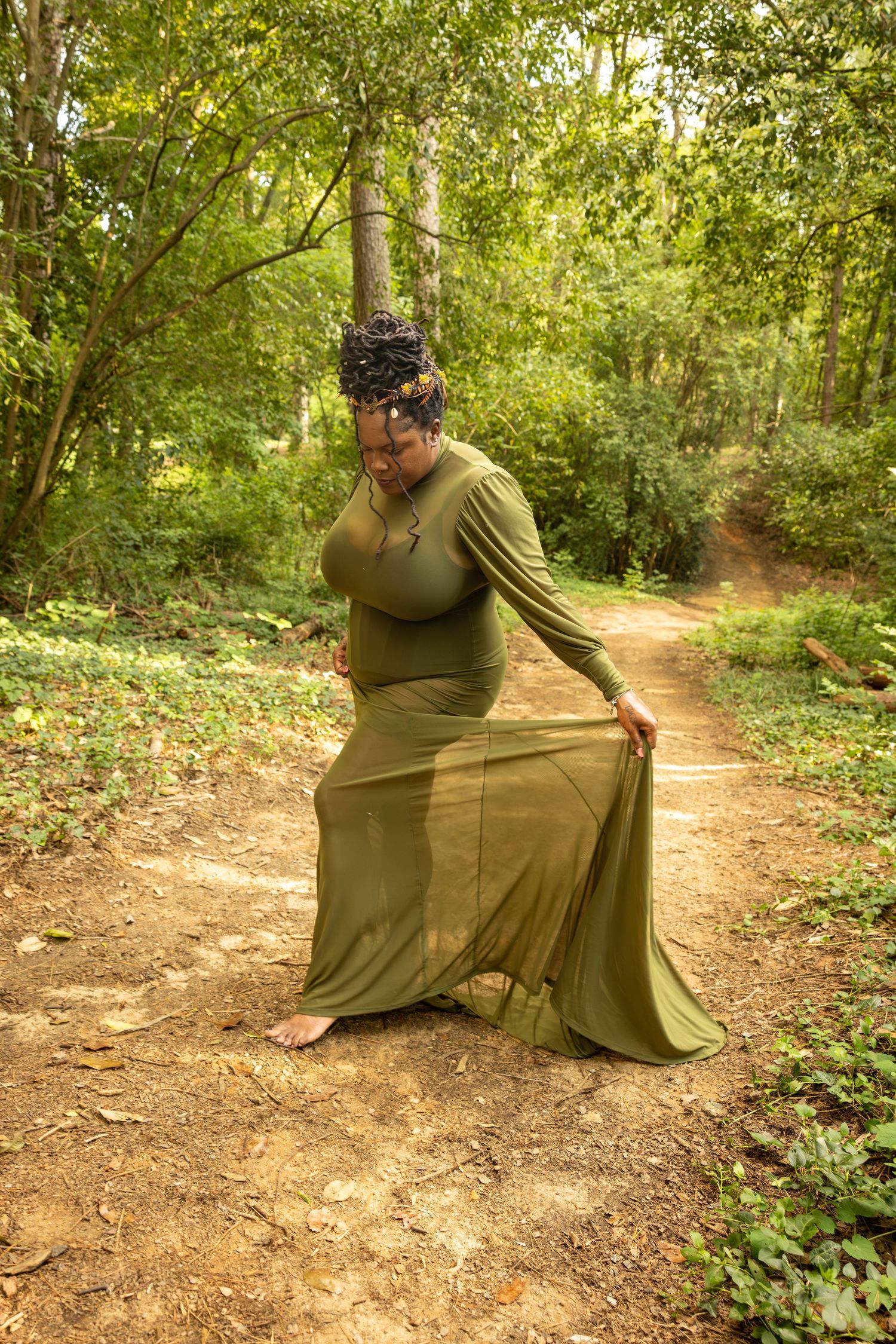 A Black woman in a flowing olive green maxi dress poses barefoot on a forest path surrounded by lush green trees.