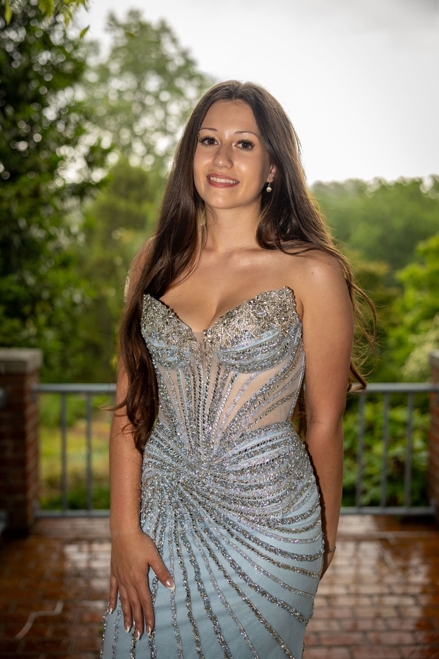 Young woman in a silver beaded strapless gown smiling on an outdoor balcony with lush greenery.