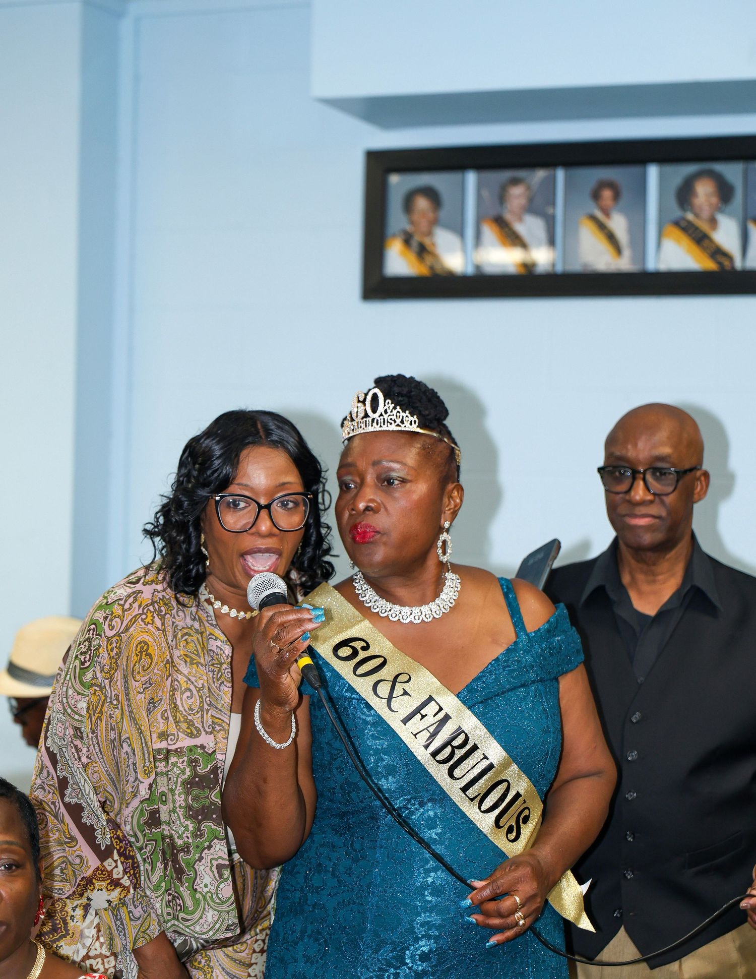 Three people at a celebration with one speaking into a microphone while wearing a sash and tiara.