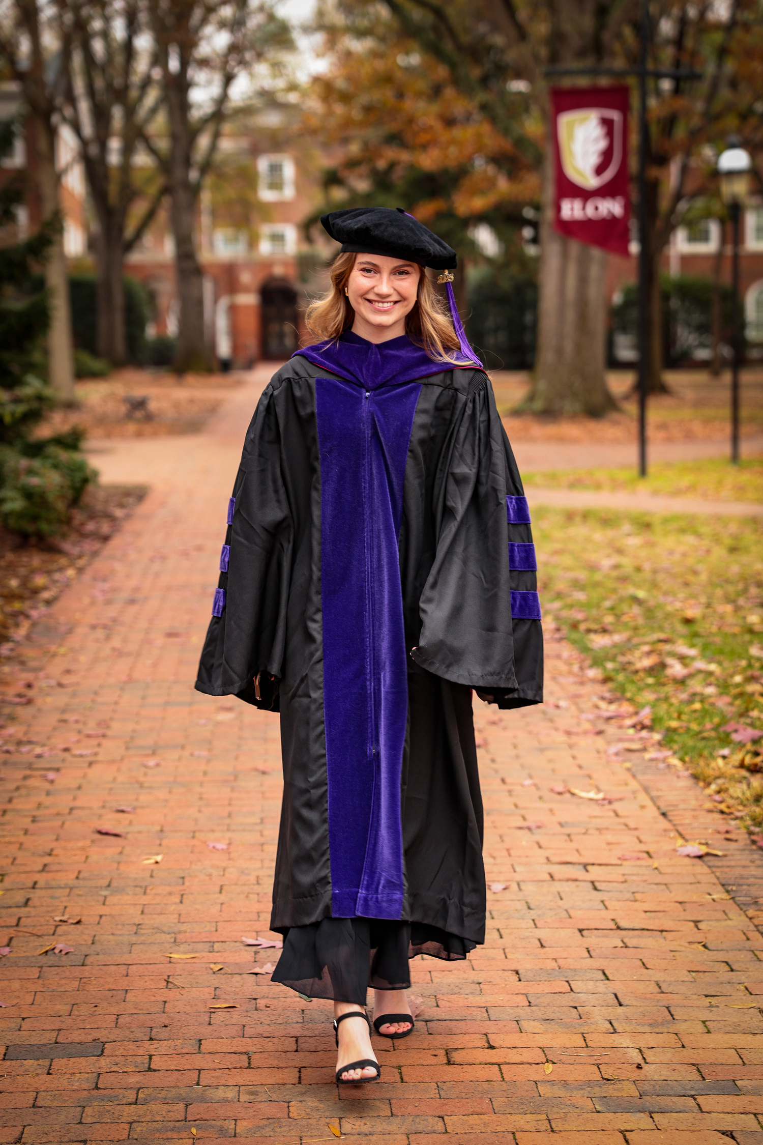 Graduate in black and purple academic regalia walks along brick pathway with university banner in background.