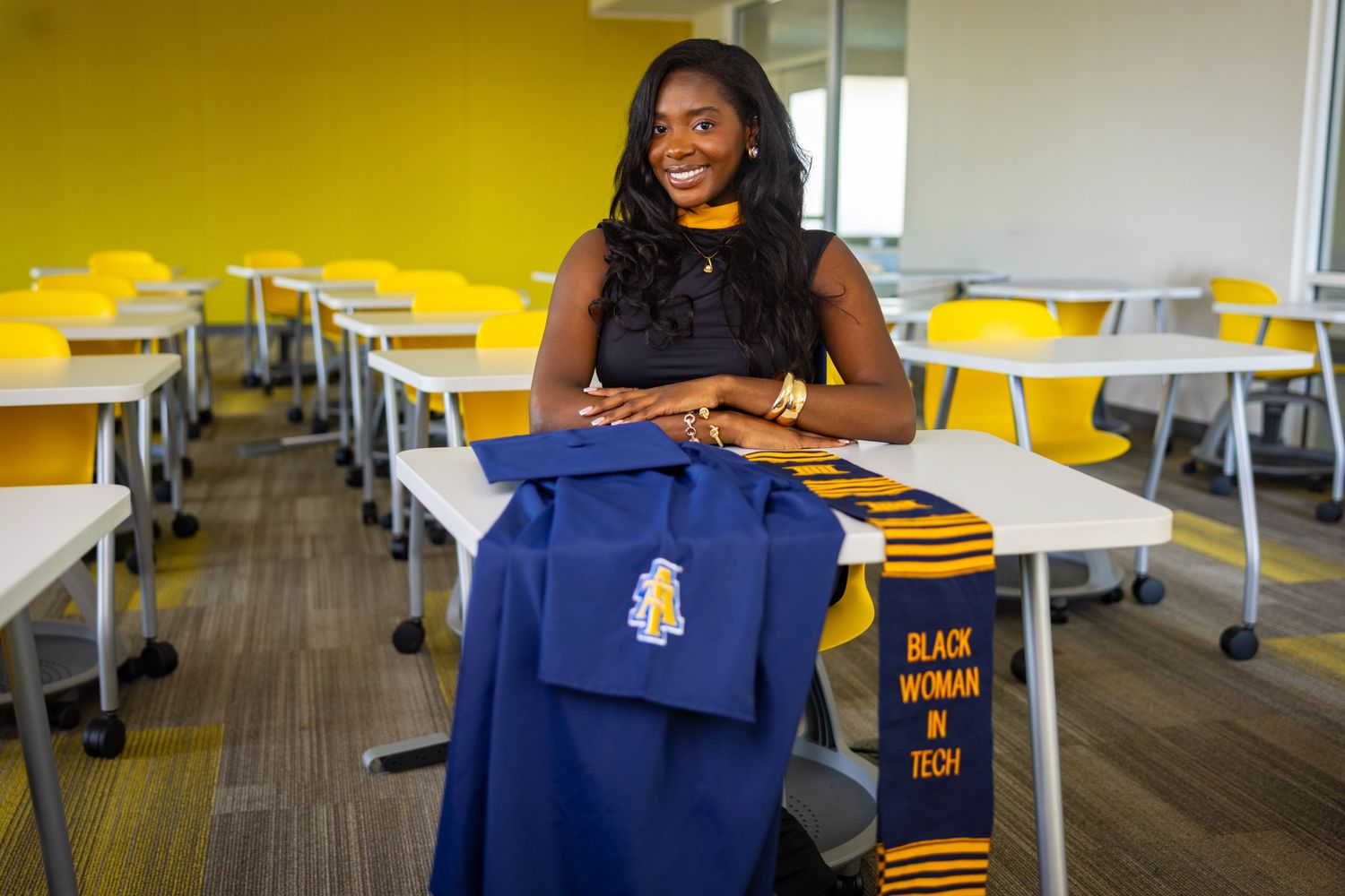 A smiling Black woman in a classroom holds blue graduation regalia with a kente stole reading Black Woman in Tech.