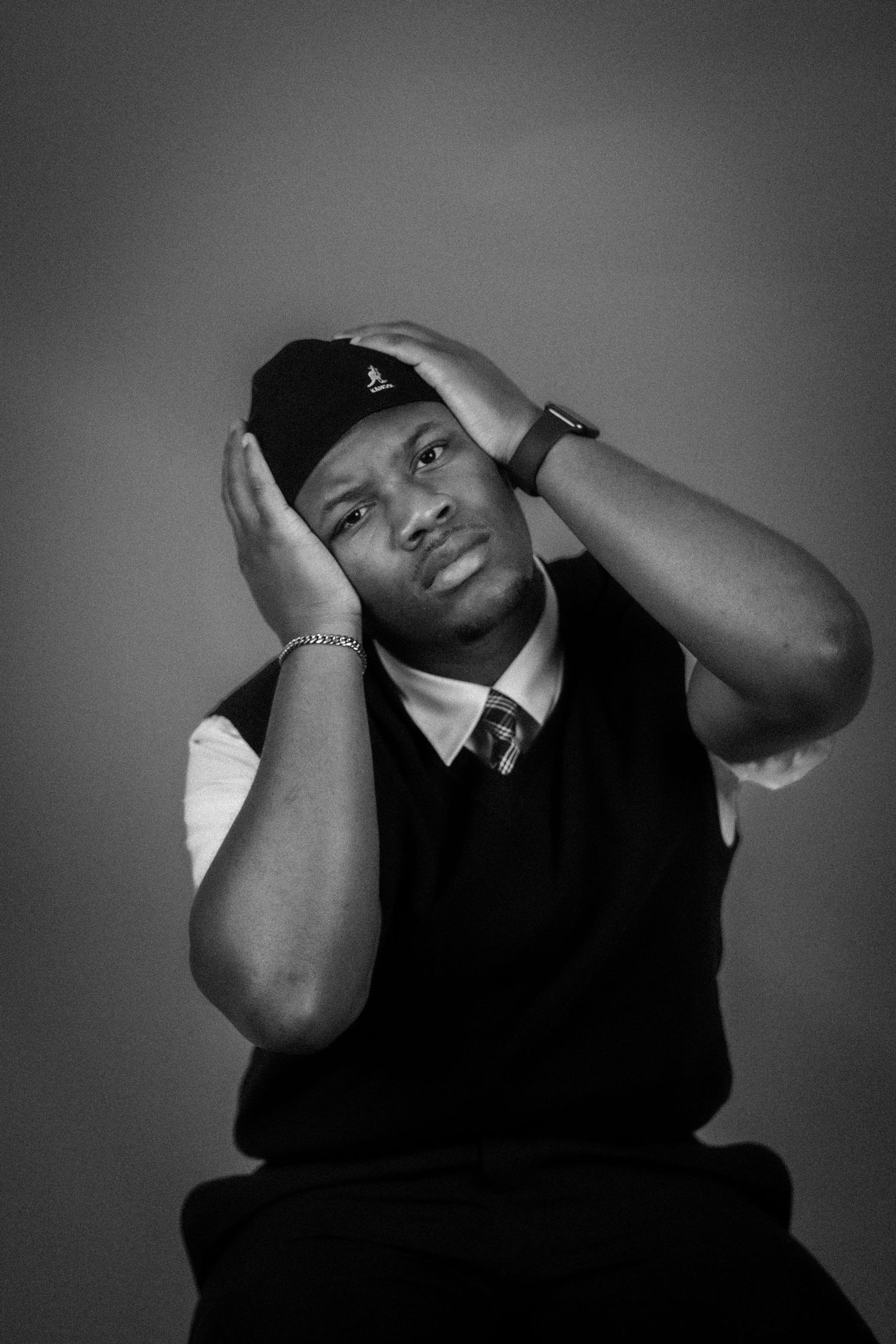Black and white portrait of a young man wearing an Adidas cap, hands on head, looking upward thoughtfully.