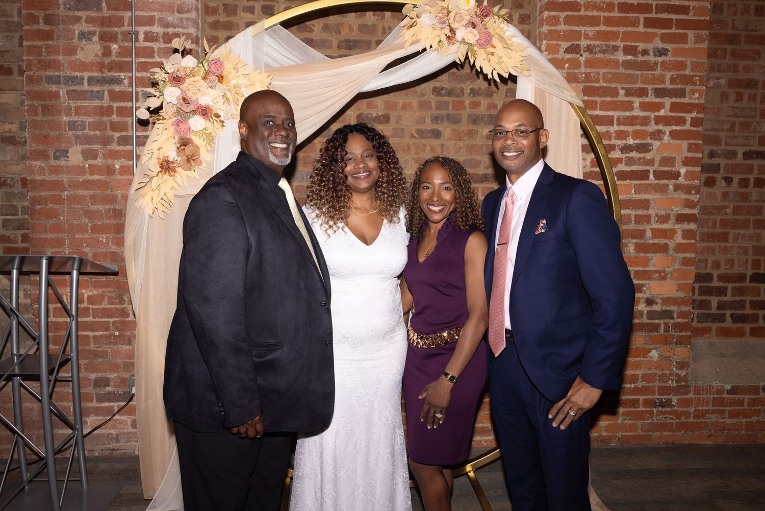 Four people smiling together under a floral arch at a wedding celebration in a brick-walled venue.