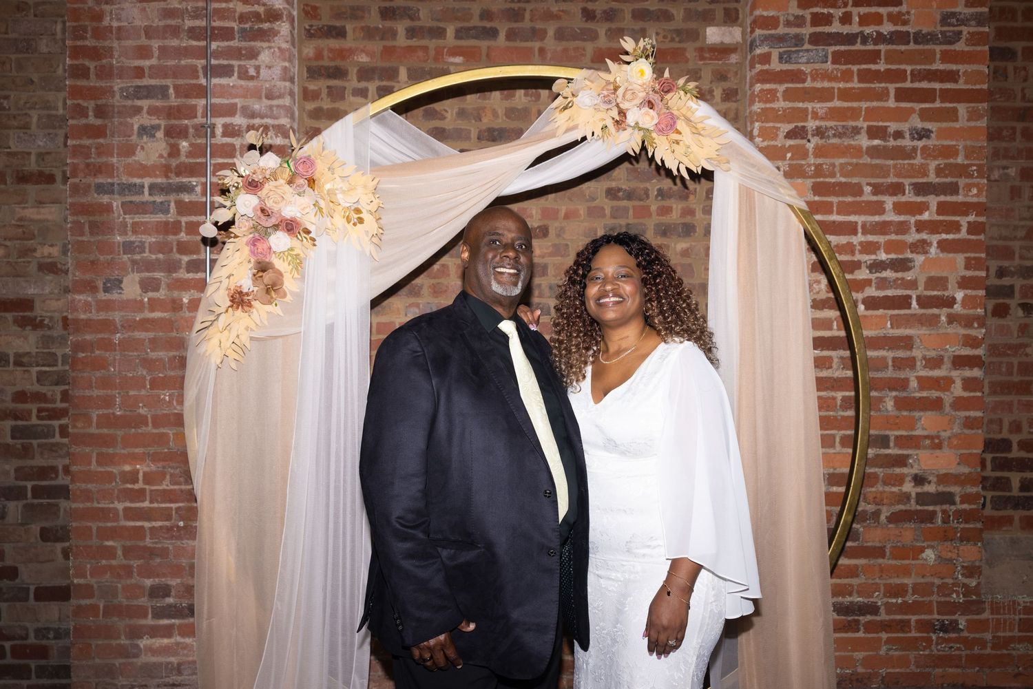 Couple posing under a gold floral arch at their wedding in a rustic brick-walled venue.