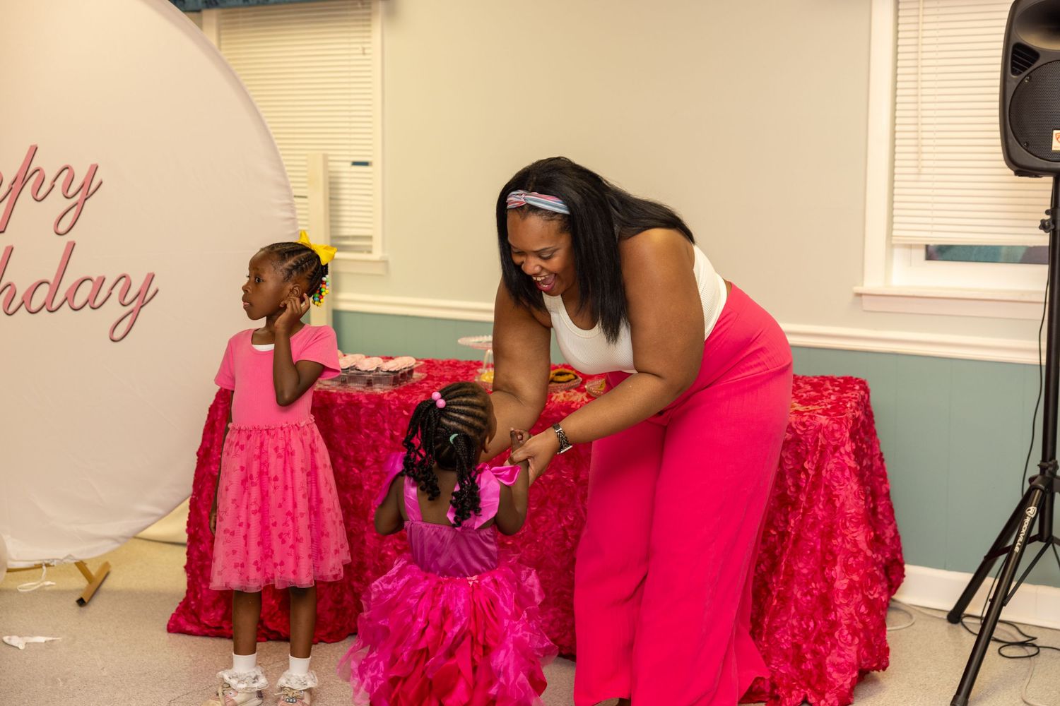 Woman in pink dress helping two young girls in pink dresses at a birthday party celebration.
