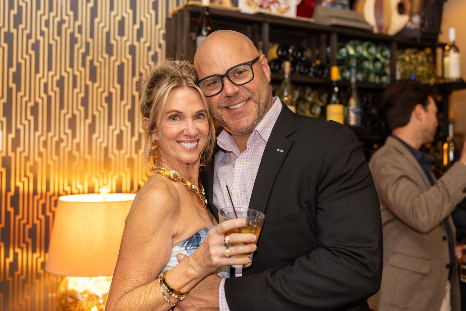 A smiling couple poses together at a bar, man holding a drink, woman in a floral top, warm ambient lighting.